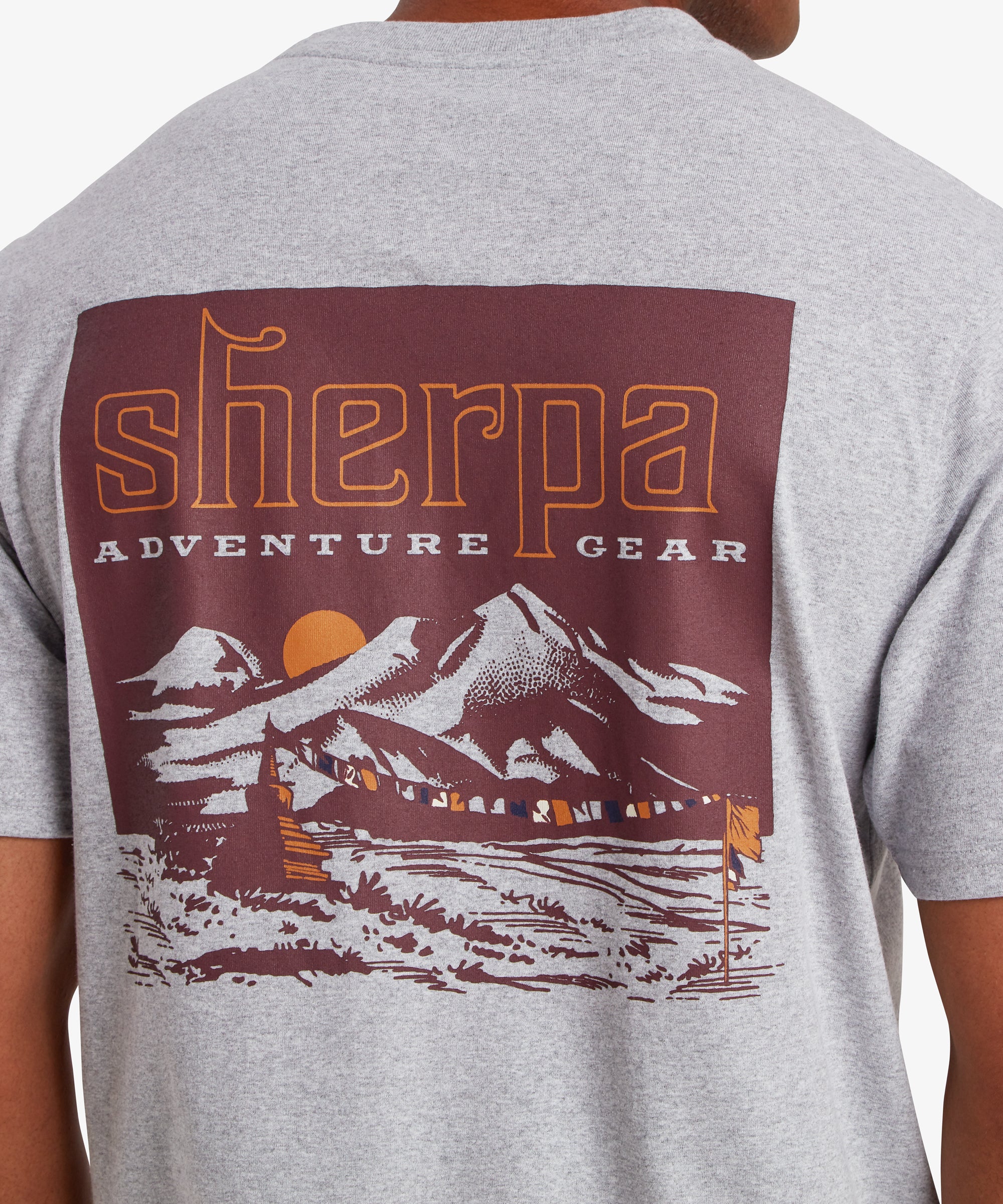 A close up back view of a man wearing a grey T-shirt with a red graphic. The printed graphic depicts a Nepal mountain range with a orange setting sun in the background and a string of multi-colored prayer flags in the foreground attached to a flag pole and small mountain shrine. The words "Sherpa Adventure Gear" sit above the mountain in orange and white.