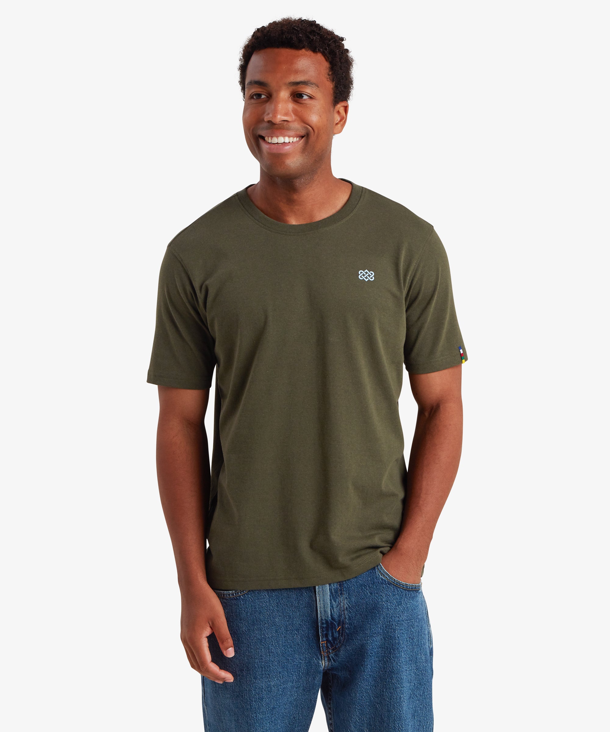 A man wearing a green T-shirt against a plain white background. The T-shirt features a small, pale blue endless knot graphic on the left chest. The T-shirt is styled with a pair of blue jeans. The man is smiling and with his left hand in his trouser pocket.