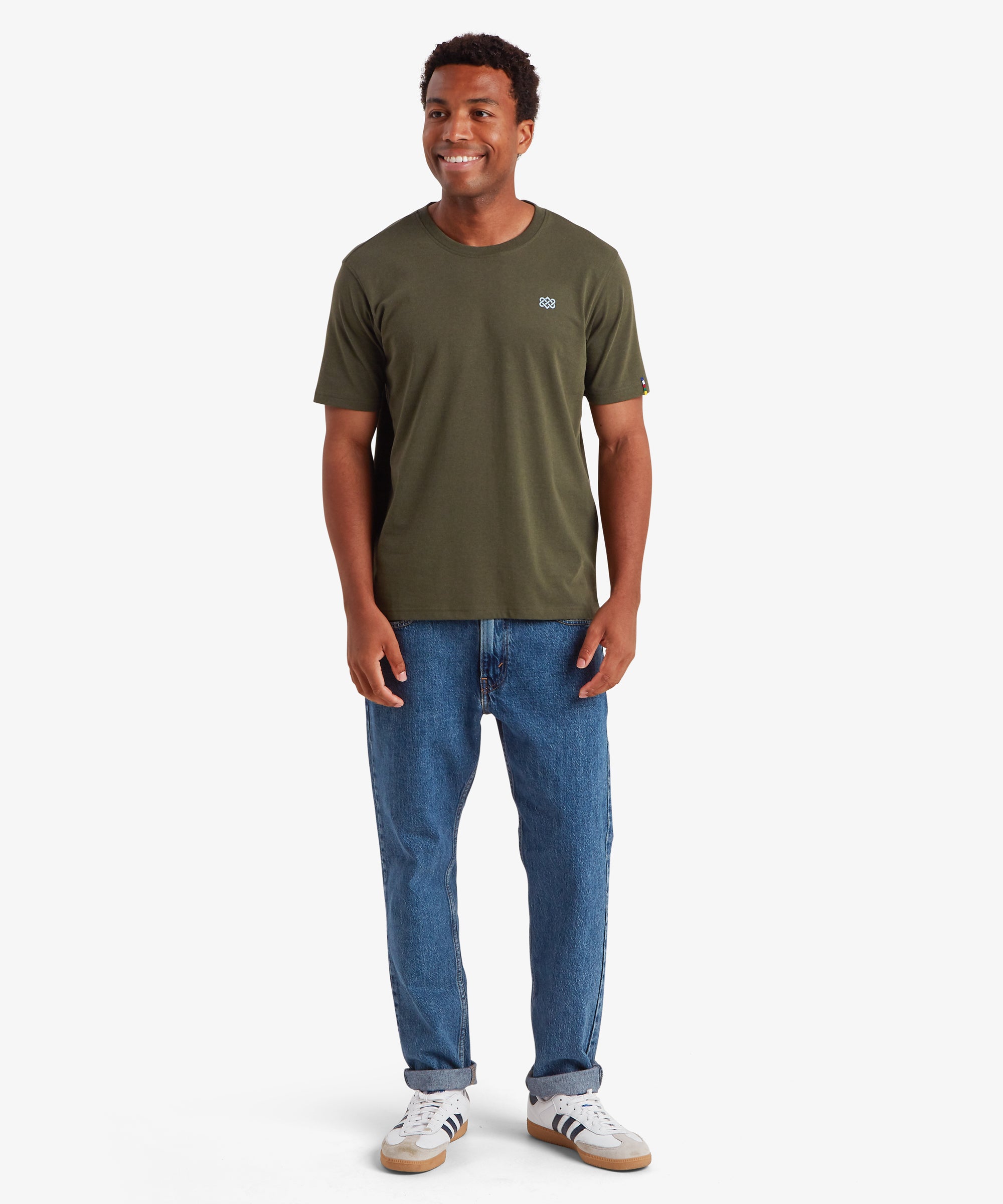 A man standing against a white background wearing a green T-shirt, blue jeans and white trainers. The T-shirt features a small, pale blue endless knot graphic on the left chest. The man is smiling and standing with his hands down by his sides.