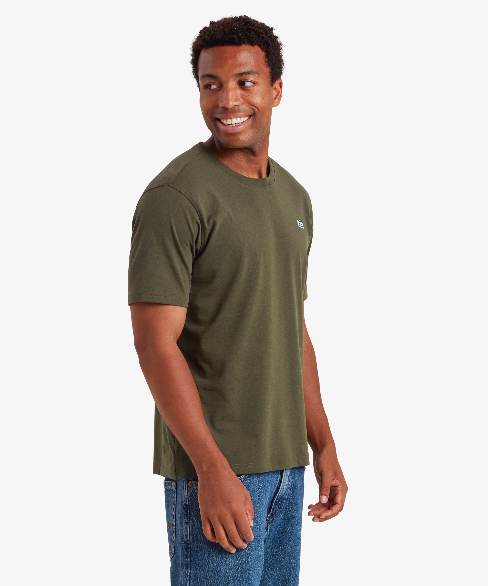 A man wearing a green T-shirt with a small blue Nepal endless knot printed on the chest. The man is pivoted to the left with both arms down by his sides. The T-shirt is styled with a pair of blue jeans creating a casual outfit for everyday wear.