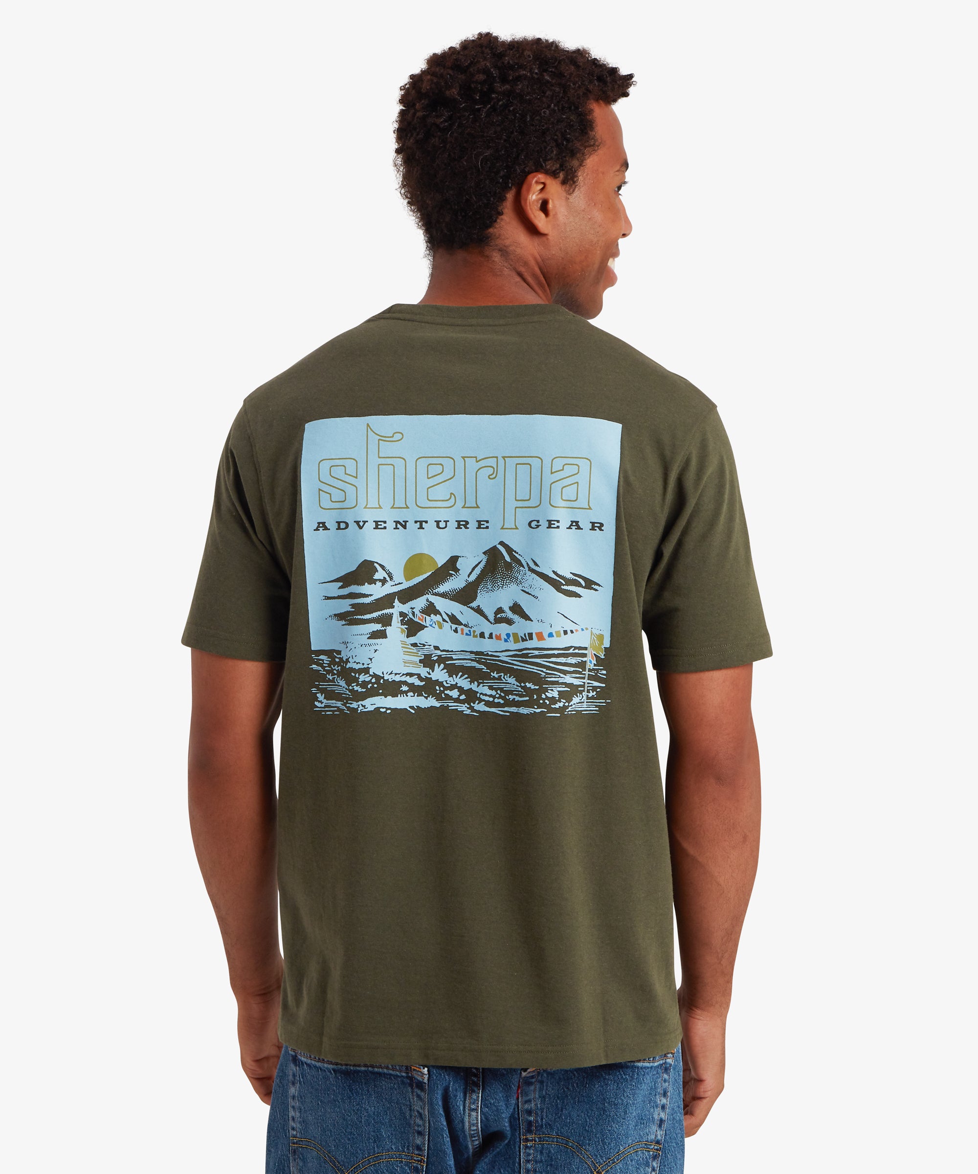 A back view of a  man wearing a short sleeve T-shirt in green with a graphic printed on the back. The graphic depicts a blue himalayan mountain range with a yellow setting sun in the background and a string of multi-colored prayer flags in the foreground attached to a flag pole and small mountain shrine. The T-shirt is styled with a pair of navy blue jeans with two pockets on the back. The man is looking slightly to the right smiling.