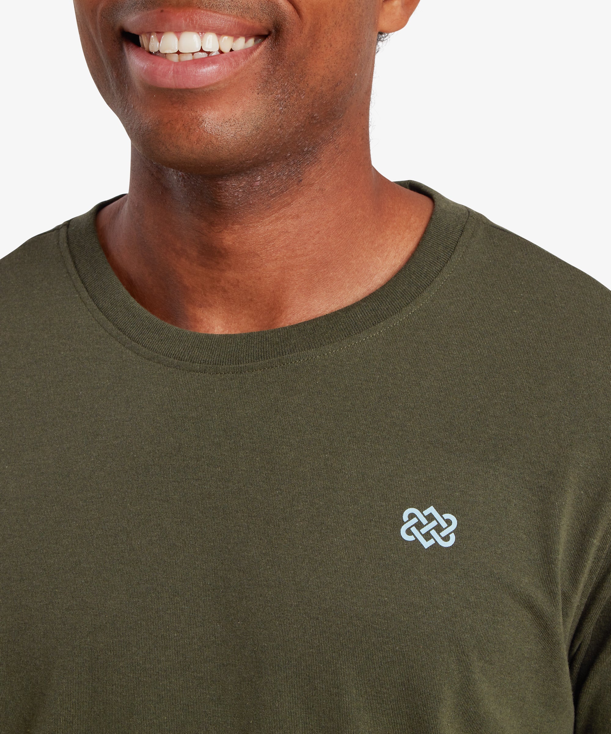 A close up of a man wearing a green T-shirt with a small pale blue Nepalese Endless knot printed on the left chest. The man is standing against a white background and smiling.