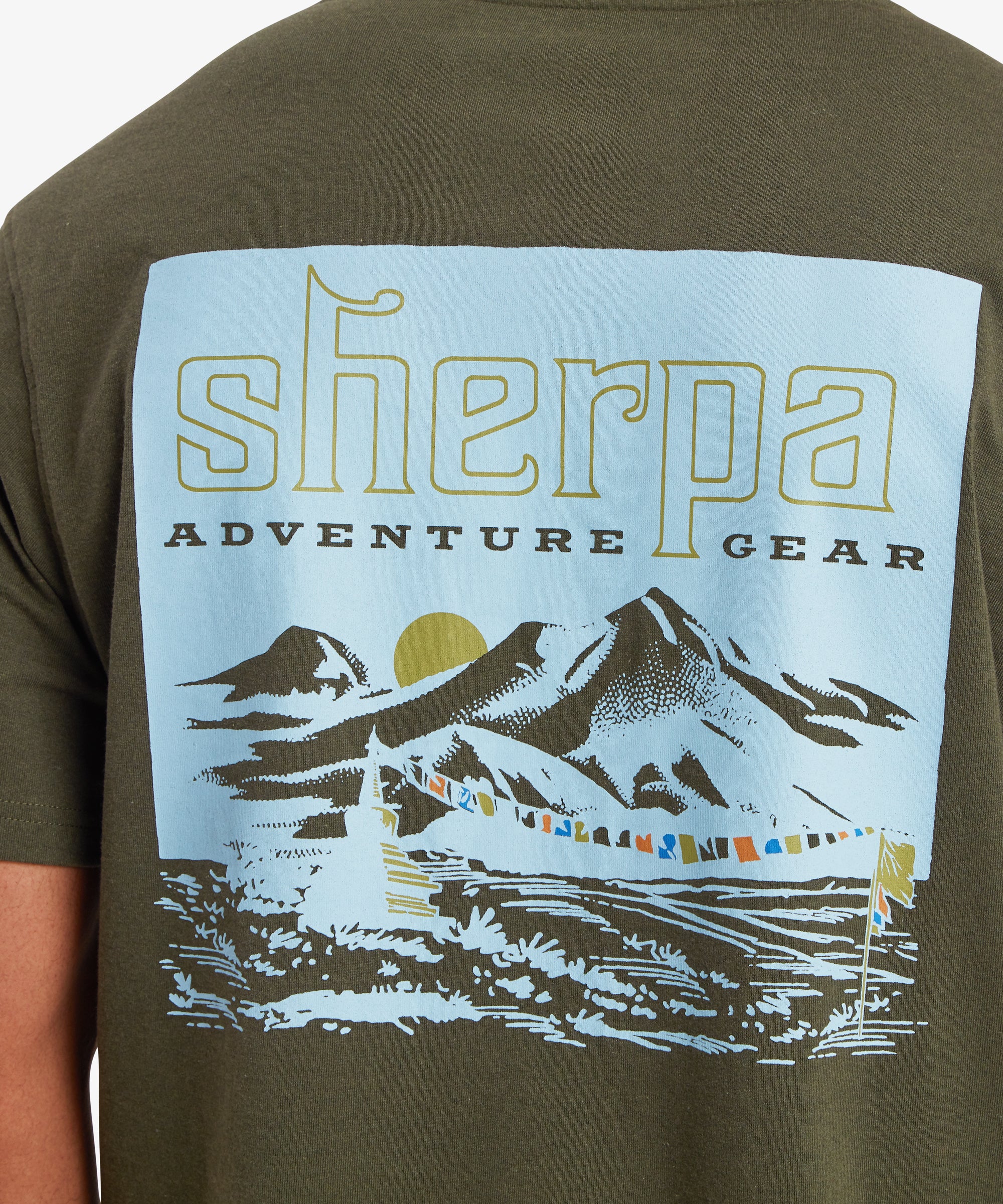 A close up of a man wearing a green T-shirt with a blue graphic printed on the back. The graphic depicts a Nepal mountain range with a yellow setting sun in the background and a string of multi-colored prayer flags in the foreground attached to a flag pole and small mountain shrine.