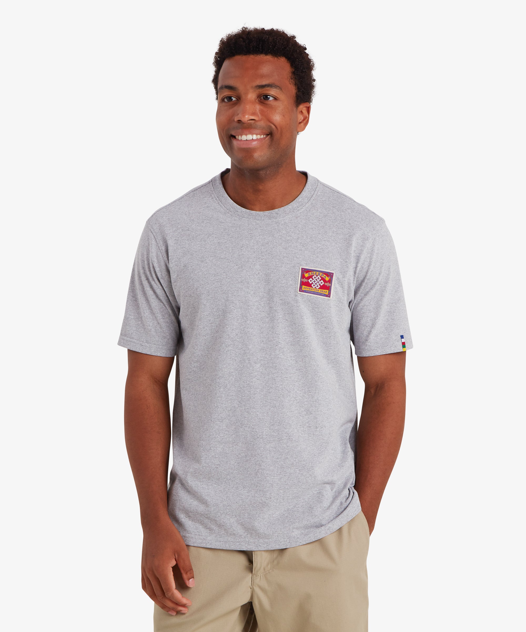 Man wearing a plain short sleeve t-shirt in grey. The T-shirt features a red patch stitched to the left chest and the word "Sherpa Adventure Gear" are visible. The patch is inspired by traditional mountaineering patches. The T-shirt also features a colored pip on the left sleeve in the 5 colors of the Nepalese prayer flags. The T-shirt is paired with beige shorts creating a relaxed everyday wear outfit ideal for summer. The man is looking slightly to the right and smiling with his left hand in his pocket.
