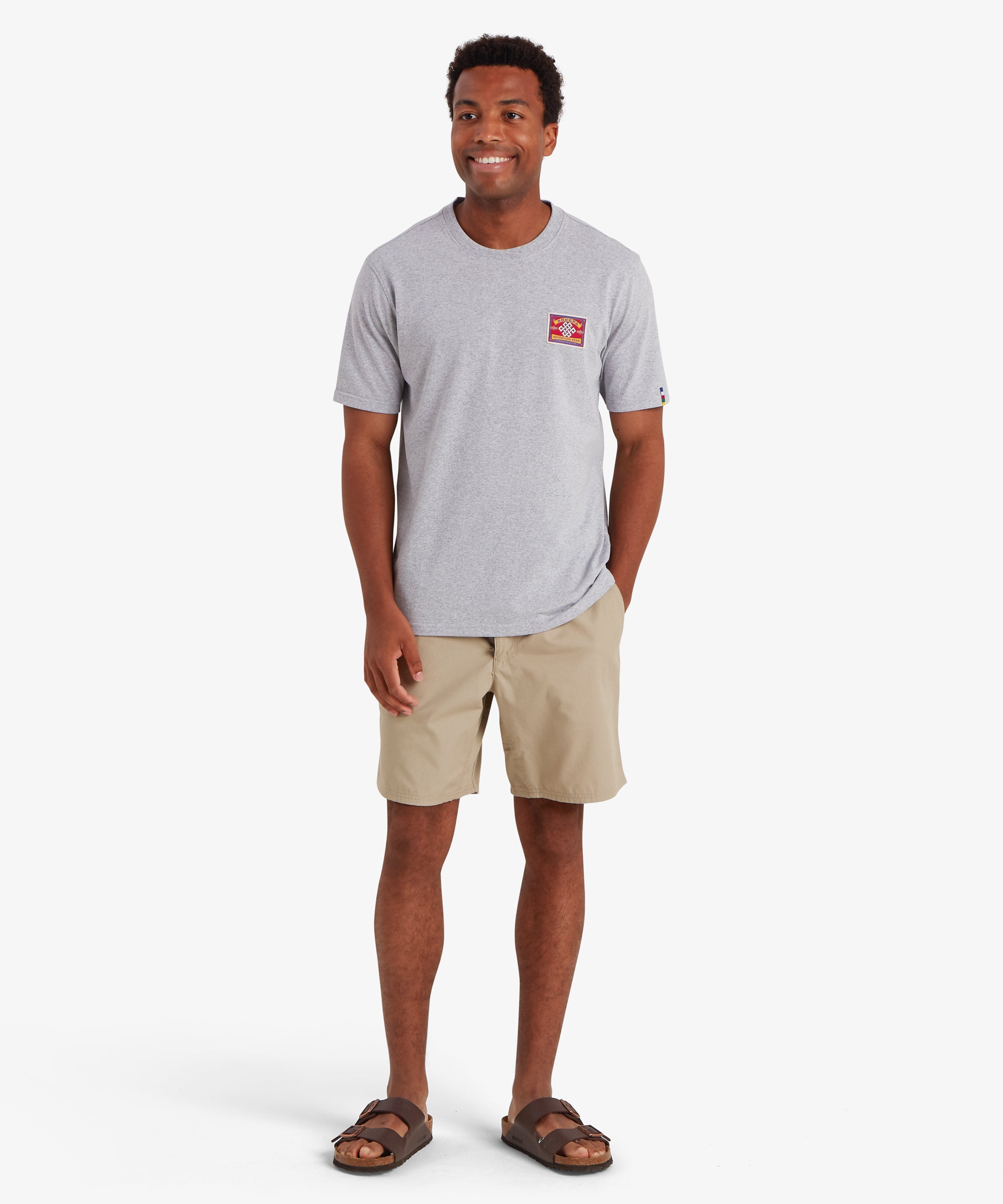 Man wearing a plain grey short sleeve t-shirt with a rectangular red patch stitched to the left chest. The patch is inspired by traditional mountaineering patches. The t-shirt also features a colored pip on the left sleeve in the 5 colors of the Nepalese prayer flags:  The T-shirt is styled with a pair of above the knee beige shorts with brown sandals creating a causal summer outfit. The man is standing looking slightly to his right with his left hand in his back pocket.