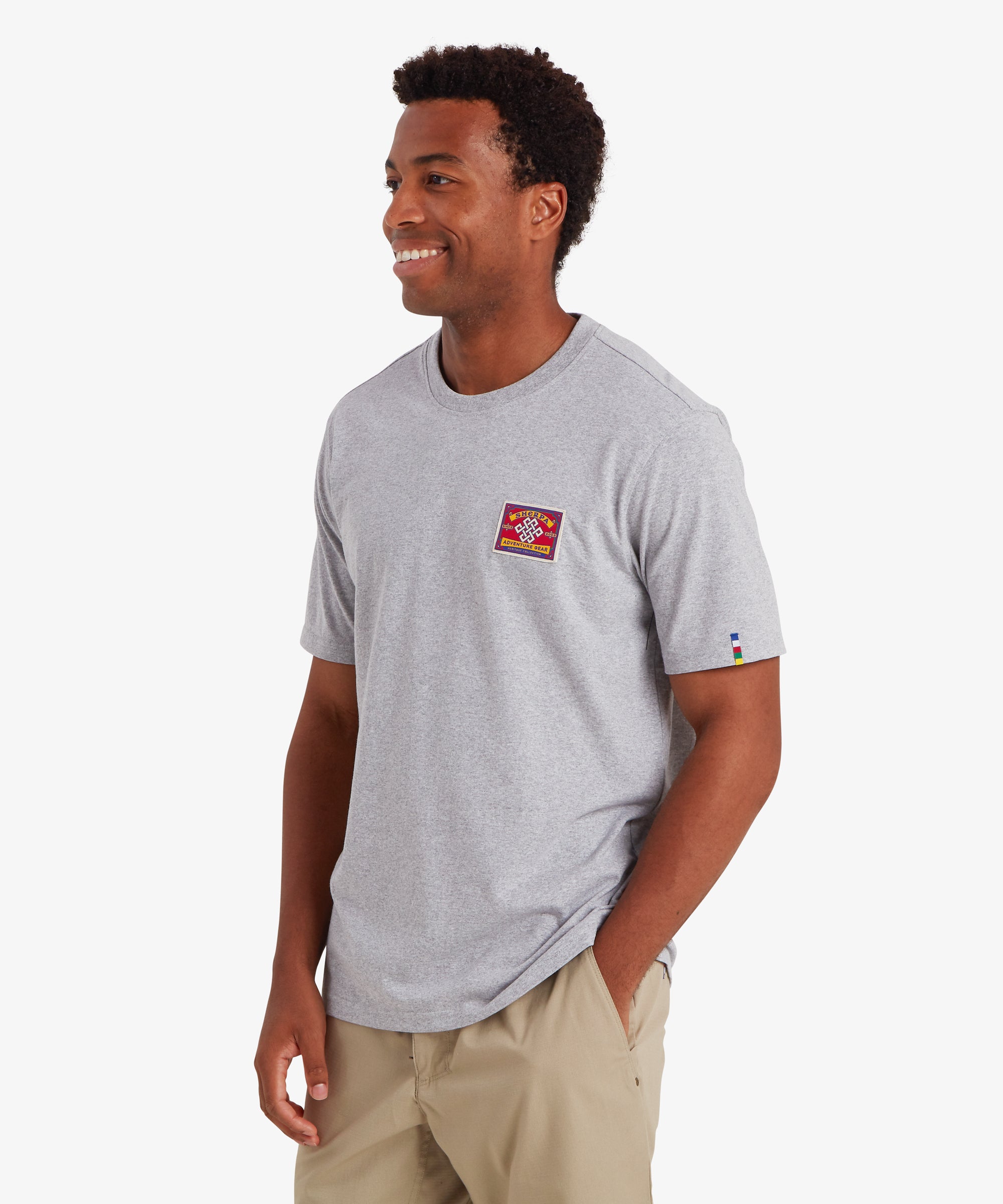 A man wearing a plain short sleeve t-shirt in grey. The T-shirt features a red patch stitched to the left chest inspired by traditional mountaineering patches. The words "Sherpa Adventure Gear" are visible on the patch. The T-shirt also features a colored pip on the left sleeve in the 5 colors of the Nepalese prayer flags. The T-shirt is paired with beige shorts creating a relaxed summer. The man is pivoted to the right with his left hand in his shorts pocket.