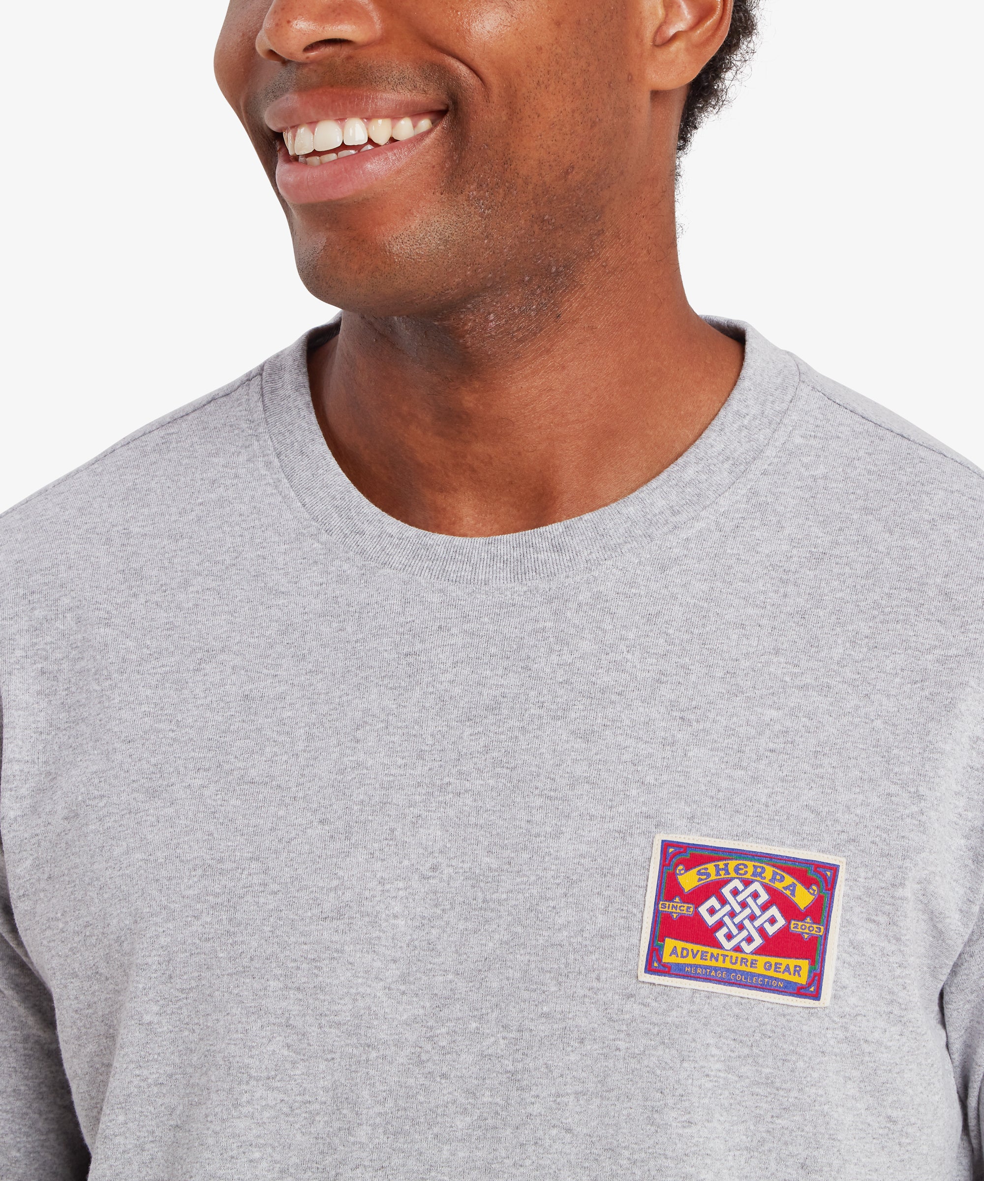 Nahaufnahme eines Mannes, der ein graues T-Shirt mit einem roten, auf die linke Brust genŠhten AufnŠher trŠgt. Der rechteckige AufnŠher hat einen wei§en Rand und die Worte ãSherpa Adventure GearÓ oben und unten. In der Mitte des AufnŠhers befindet sich ein wei§er Endlosknoten. Der Mann steht lŠchelnd vor einem wei§en Hintergrund.