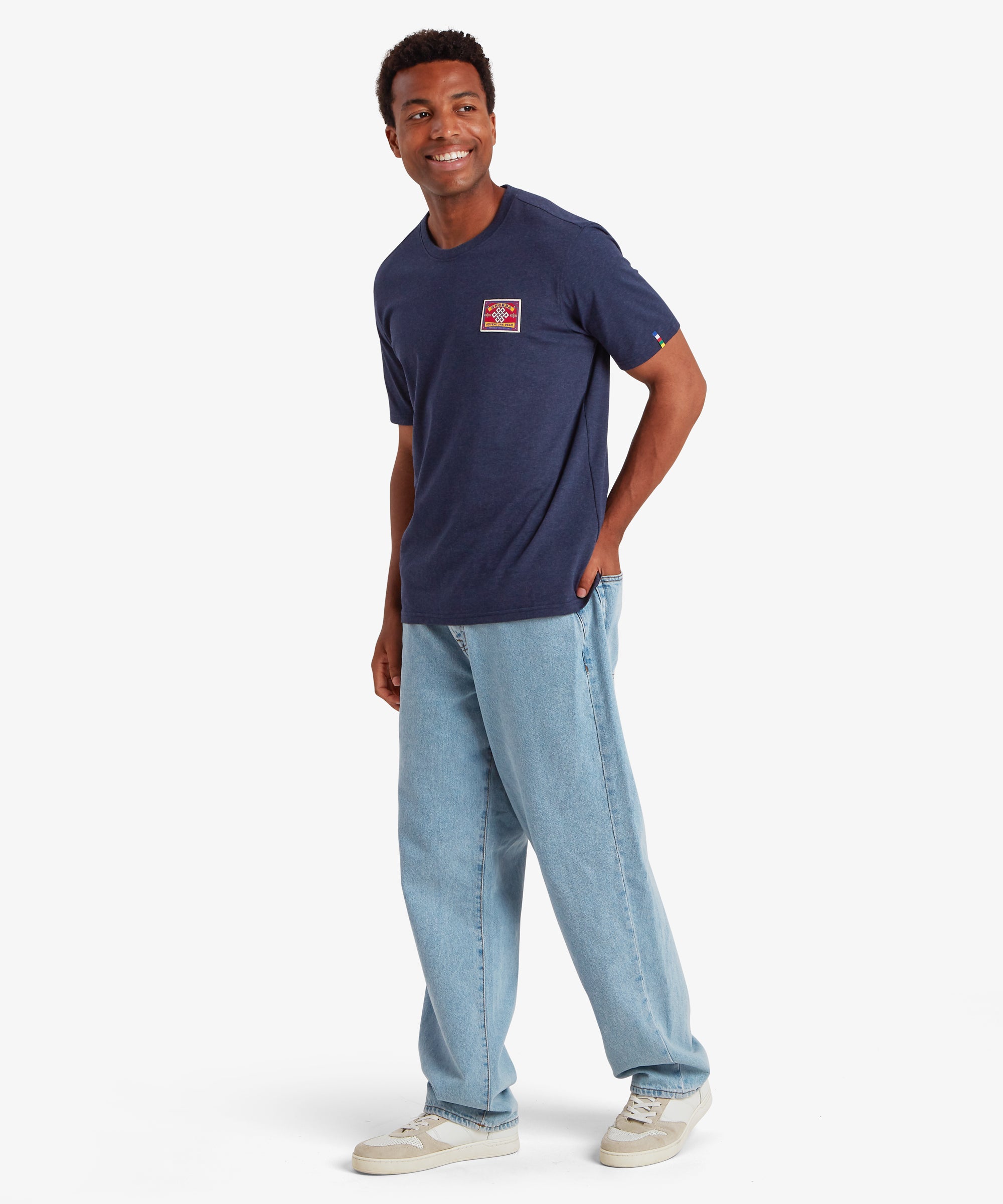 Man wearing a navy short sleeve t-shirt with a rectangular red patch on the left chest. The patch is inspired by traditional mountaineering patches. The t-shirt also features a colored pip on the left sleeve in the 5 colors of the Nepalese prayer flags:  The T-shirt is styled with a pair of baggy blue jeans and white trainers creating a relaxed everyday wear outfit. The man is standing looking slightly to his right with his left hand in his back pocket.