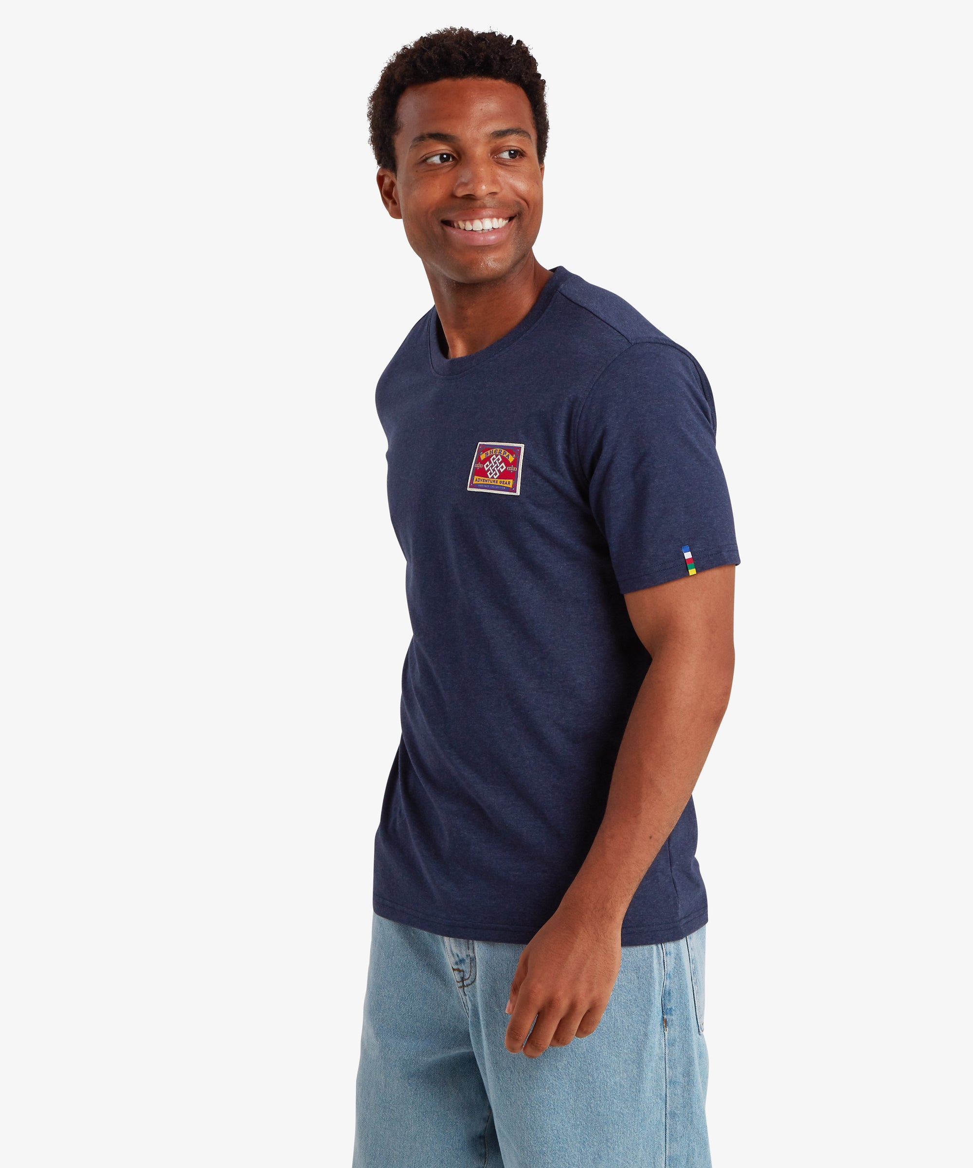 Ein Mann trŠgt ein marineblaues Kurzarm-T-Shirt mit einem rechteckigen roten AufnŠher auf der linken Brust. Der AufnŠher ist von traditionellen Bergsteiger-AufnŠhern inspiriert. Das T-Shirt hat au§erdem einen farbigen Streifen auf dem linken €rmel in den fŸnf Farben der nepalesischen Gebetsfahnen:  Das T-Shirt wird mit einer weiten blauen Jeans kombiniert, wodurch ein lŠssiges Outfit fŸr den Alltag entsteht. Der Mann ist nach rechts gedreht und hat die HŠnde an den Seiten herunterhŠngen.