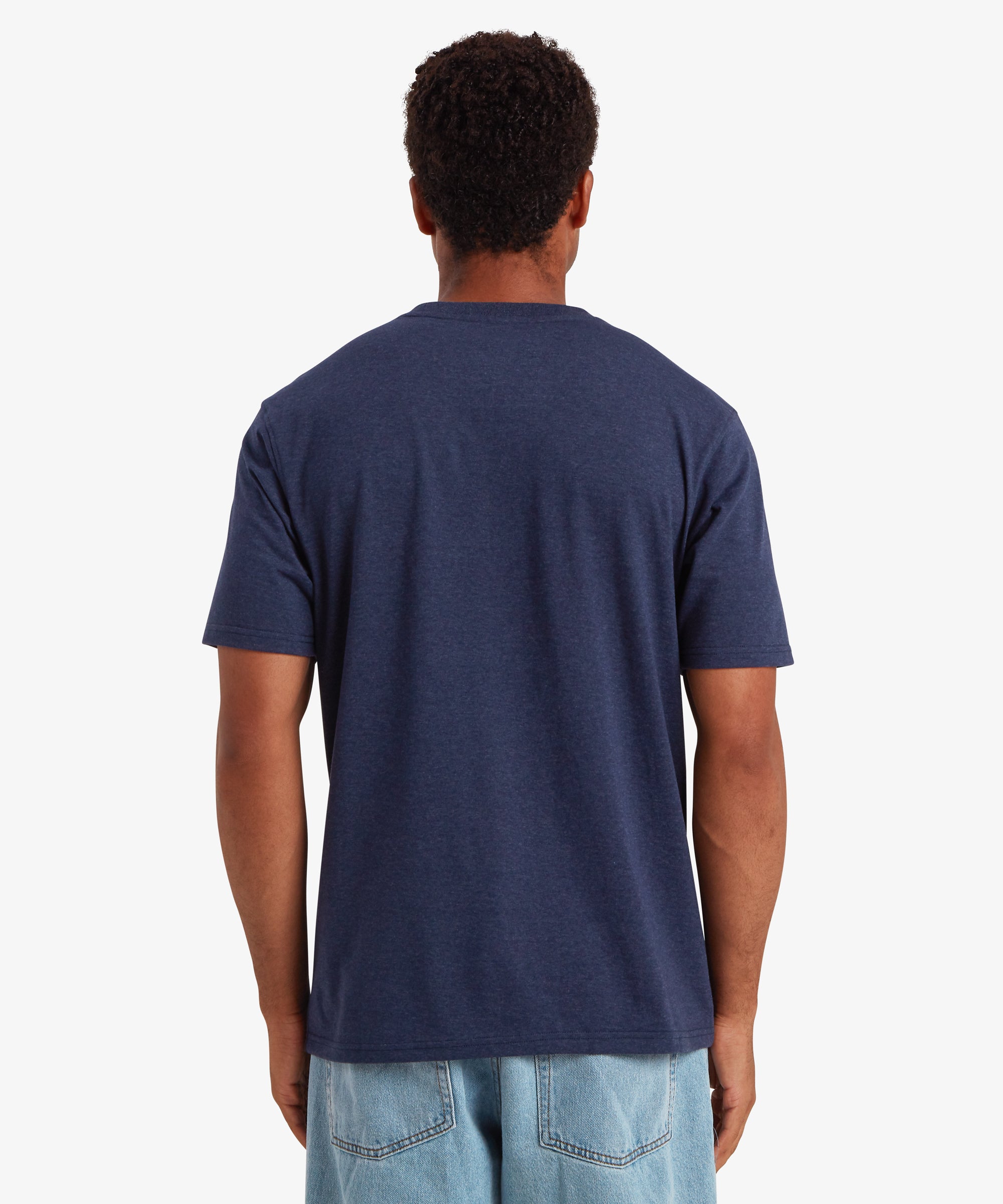 A back view of a man wearing a plain navy short sleeve t-shirt. The t-shirt is styled with a pair of loose blue jeans. The man is standing against a white background with his hands down by his sides.