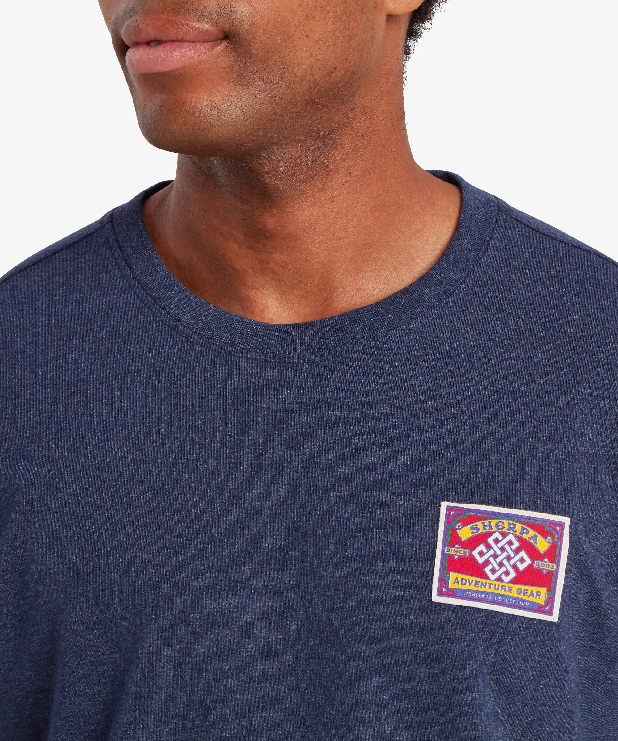 Nahaufnahme eines Mannes, der ein marineblaues T-Shirt mit einem aufgestickten Patch im Stil der nepalesischen Bergsteiger-Tradition auf der linken Brust trŠgt. Der rechteckige Patch ist rot mit wei§em Rand und der Aufschrift ãSherpa Adventure GearÓ auf gelbem Hintergrund. In der Mitte des Patches befindet sich ein wei§er Endlosknoten. Der Mann steht vor einem wei§en Hintergrund und lŠchelt leicht.