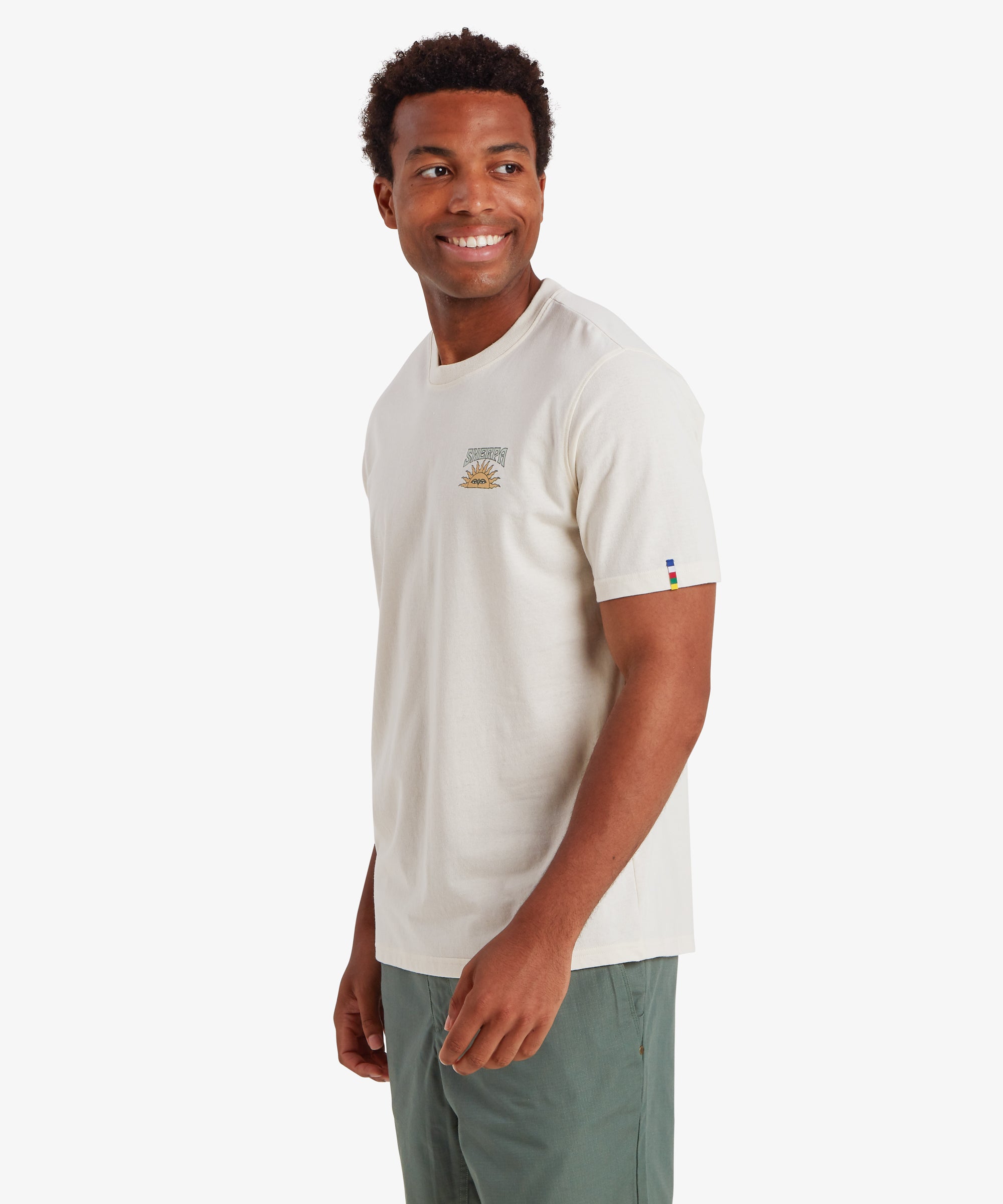 A man wearing a white short sleeve t-shirt with a small graphic logo on the left chest. The small graphic depicts a traditional Nepali setting sun with the word "Sherpa" printed above it in blue. The T-shirt also features a colored pip on the left sleeve in the 5 colors of the Nepalese prayer flags. The T-shirt is paired with green shorts creating a relaxed everyday wear outfit ideal for summer. The man's body is pivoted to the right and he is looking over his shoulder.