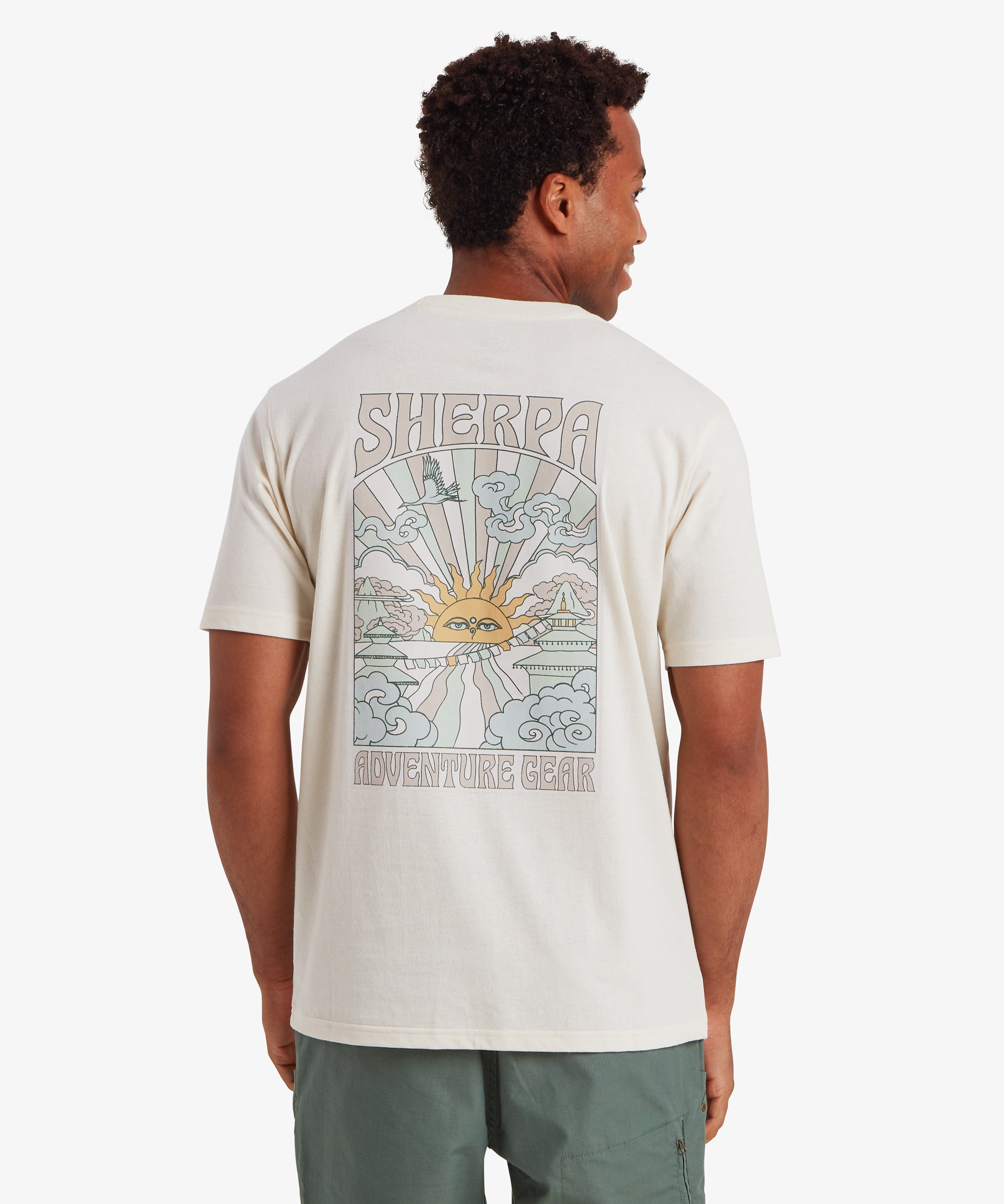A back view of a man wearing a white short sleeve T-shirt with a pale blue, pink and yellow graphic on the back. The vertical rectangular graphic depicts a traditional Nepalese sun at the centre emitting beams of pale pink and blue light. Clouds, traditional Nepali temples and other iconography sit at the adorn the edges of the graphic. The words "Sherpa" is written at the top of the graphic and "Adventure Gear" at the bottom. The T-shirt is styled with green shorts with a zipped pocket on the side.