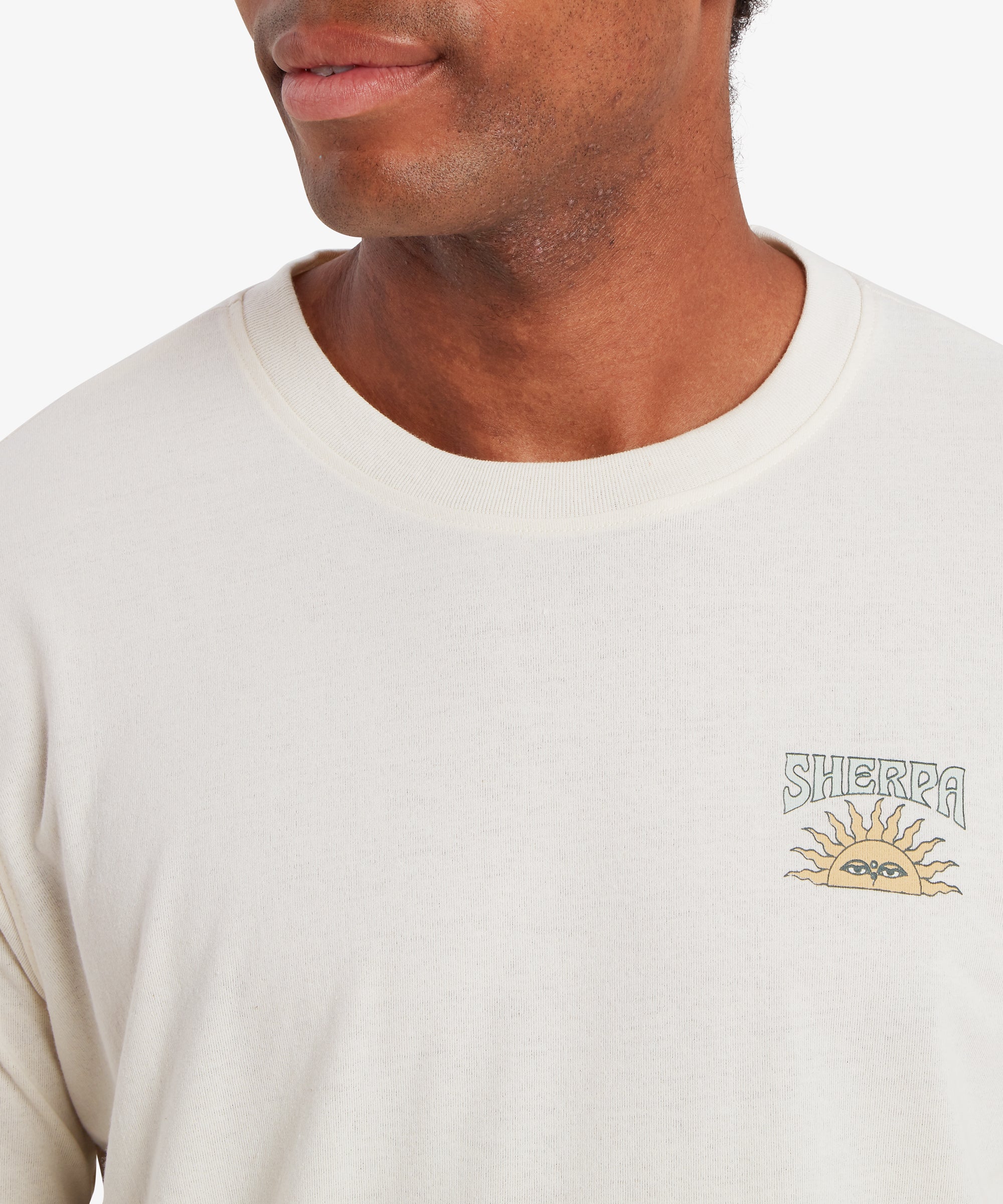 A close up of a man wearing a white T-shirt. The T-shirt features a small graphic of a traditional Nepal setting sun with the word "Sherpa" written above it in a pale blue with a solid blue outline.