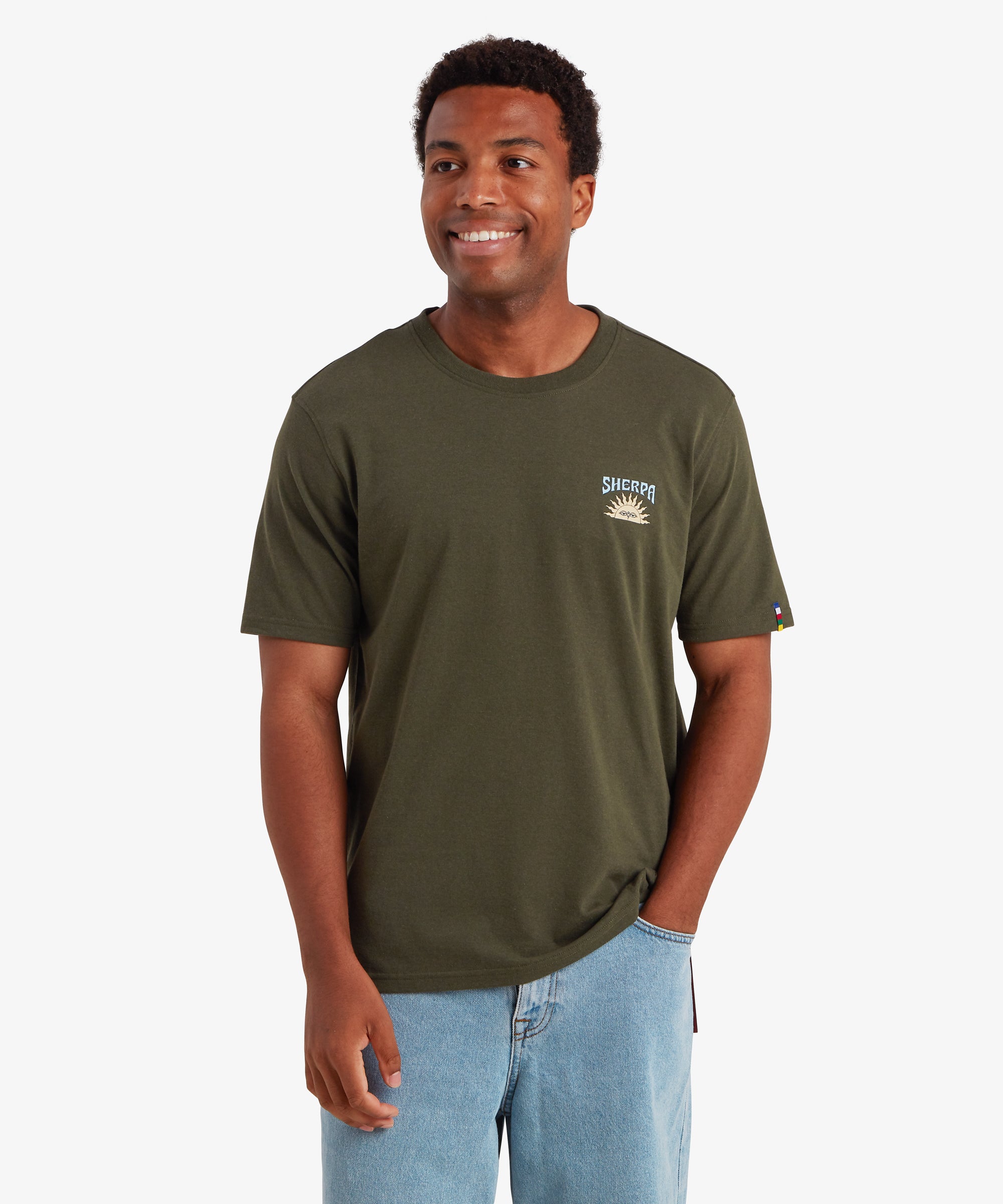 A man wearing a plain short sleeve t-shirt in green. The T-shirt features a small graphic of a traditional Nepali setting sun with the word "Sherpa" printed above it. The T-shirt also features a colored pip on the left sleeve in the 5 colors of the Nepalese prayer flags. The T-shirt is paired with pale blue jeans creating a relaxed everyday wear outfit ideal for summer. The man is looking slightly to the right and smiling with his left hand in his pocket.