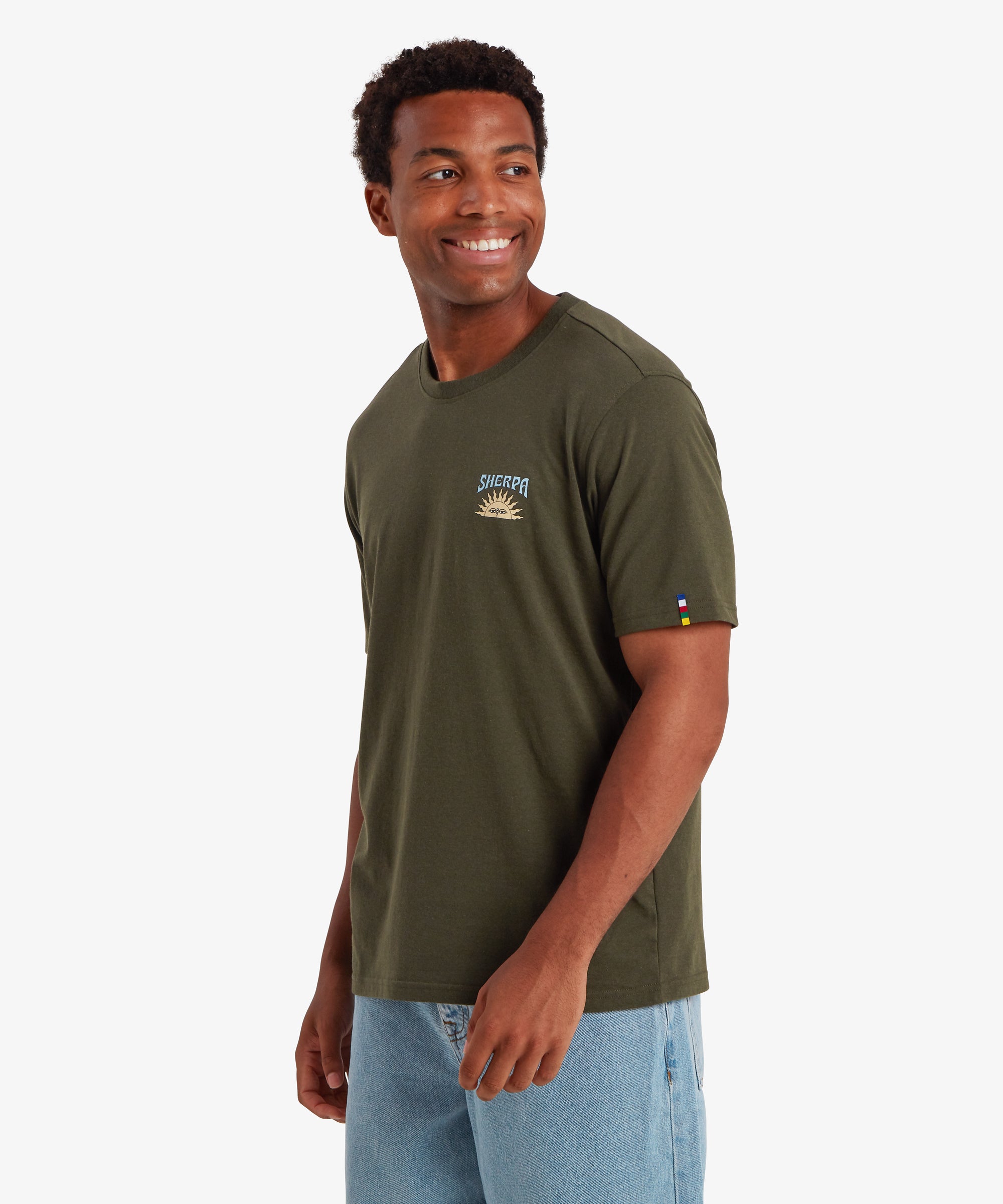 A man wearing a green short sleeve t-shirt. The T-shirt features a small graphic printed on the chest. The graphic depicts a traditional Nepali setting sun with the word "Sherpa" printed above it. The T-shirt also features a colored pip on the left sleeve in the 5 colors of the Nepalese prayer flags. The T-shirt is paired with loose pale blue jeans creating a relaxed everyday wear outfit. The man's body is pivoted to the right and he is looking over his shoulder smiling with both hands down by his sides.