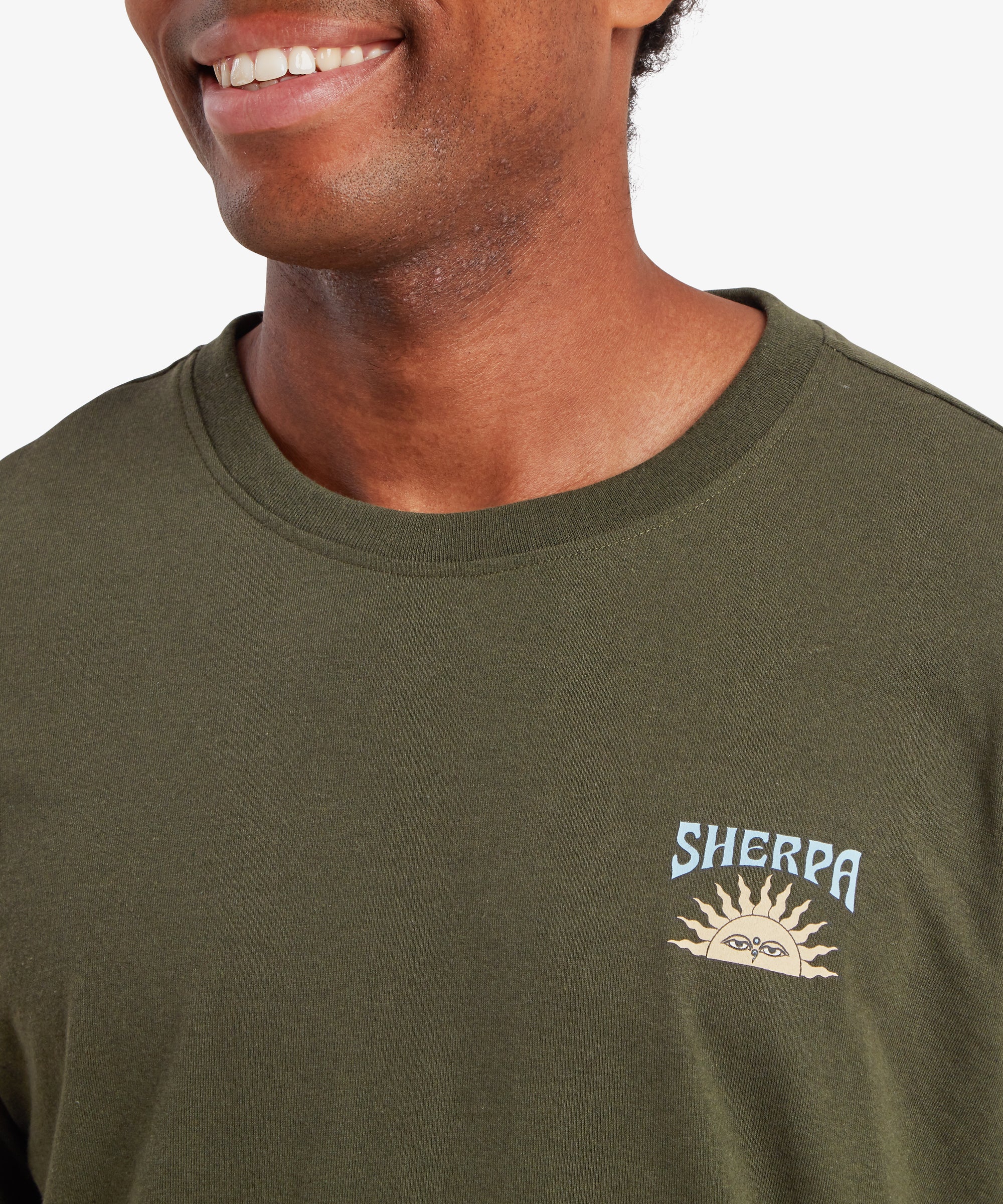 A close up of a man wearing a green T-shirt. The T-shirt features a small graphic of a traditional Nepal setting sun with the word "Sherpa" written above it.