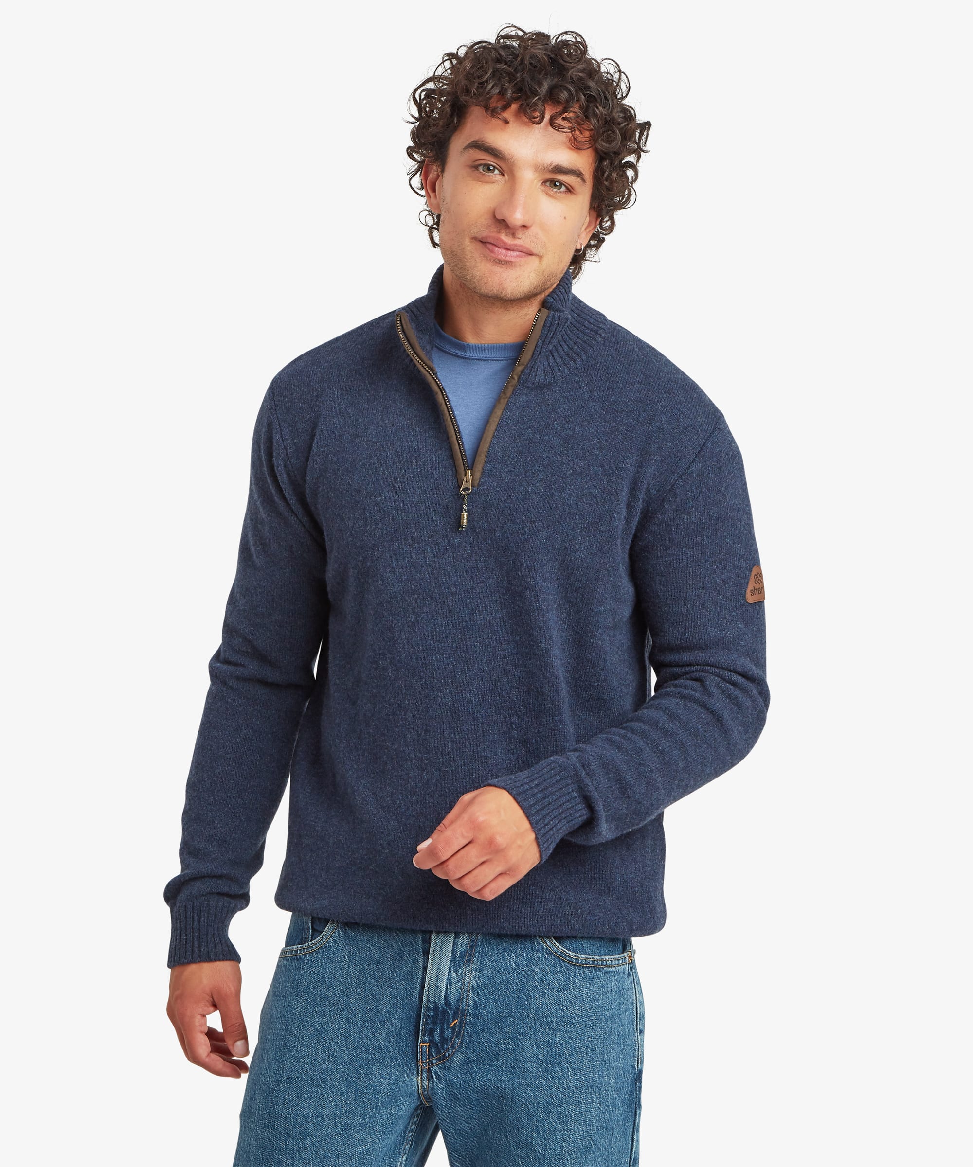 A man wearing a navy blue quarter-zip knit sweater with brown trim and a small brown Sherpa Adventure logo patch on the sleeve, paired with blue jeans, against a light gray background.