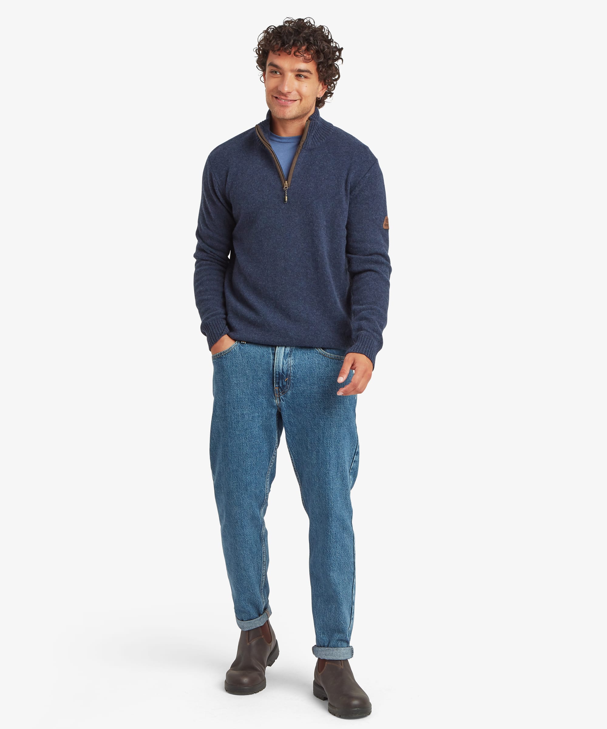 A full length portrait man modeling a navy blue quarter-zip sweater with a brown collar, paired with blue jeans and brown boots, against a light gray studio background. The sweater is finished with ribbing on the hem, collar and cuffs as well as a leather Sherpa Adventure Gear patch on the sleeve.