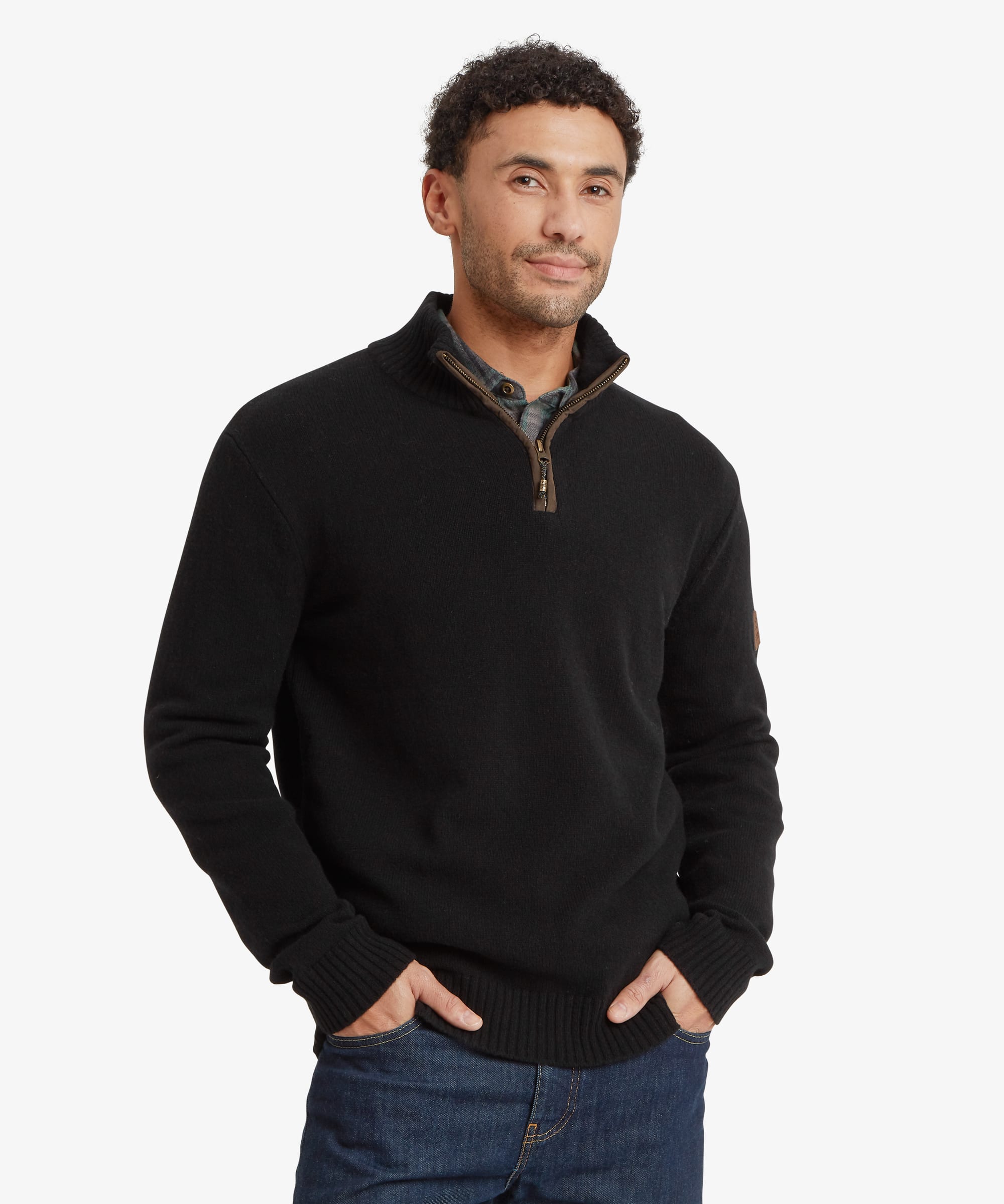 A man wearing a black quarter-zip knit sweater with a brown zipper and over a shirt with a subtle plaid collar, paired with blue jeans, against a light gray background. The sweater features leather trim on the zip and a Sherpa Adventure Gear endless knot zip puller.