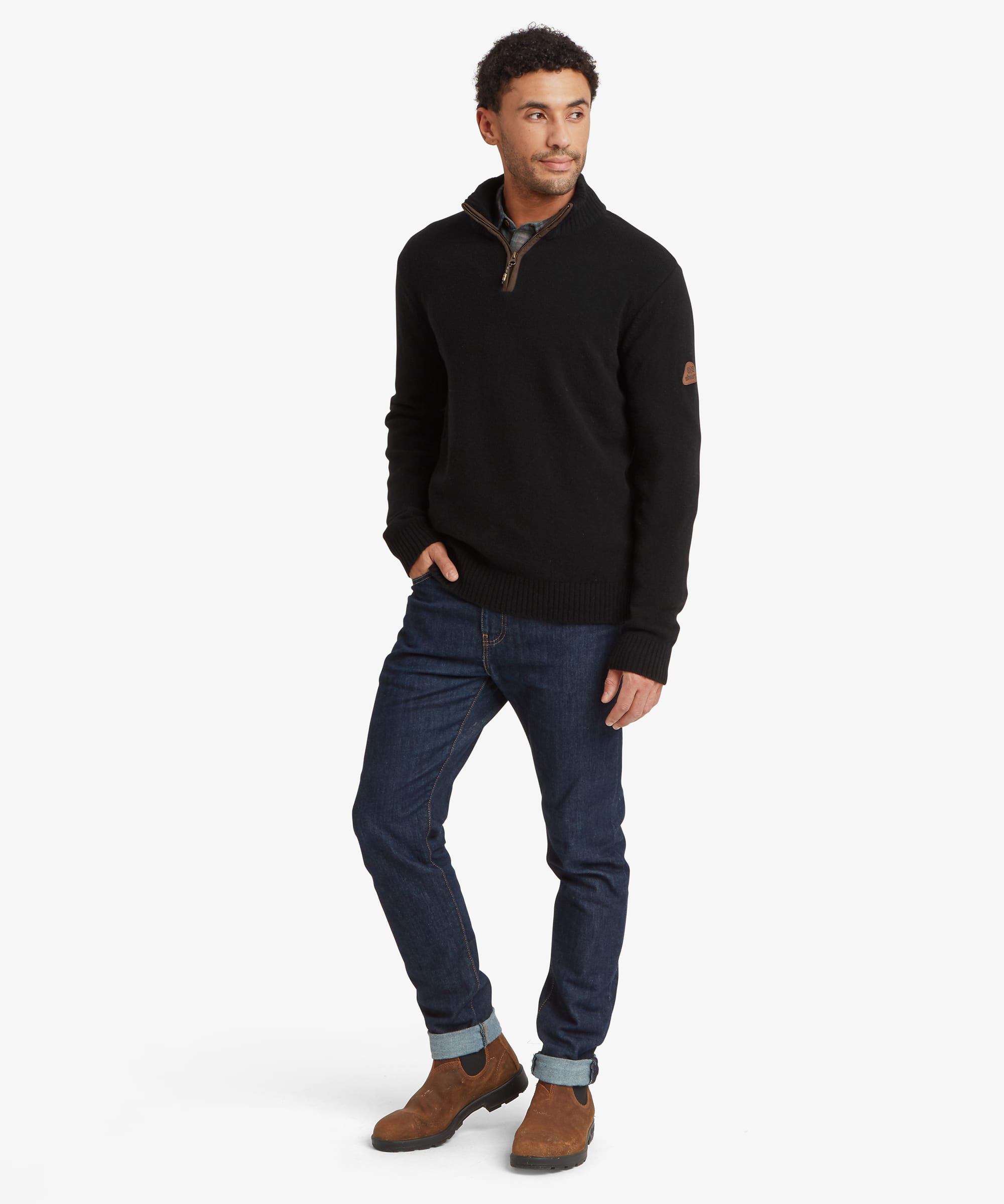 A man modeling a black quarter-zip pullover sweater with a brown sleeve patch, paired with dark blue jeans and brown suede boots, against a light gray studio background. The sweater features a leather patch on the sleeve with the Sherpa Adventure Gear logo.