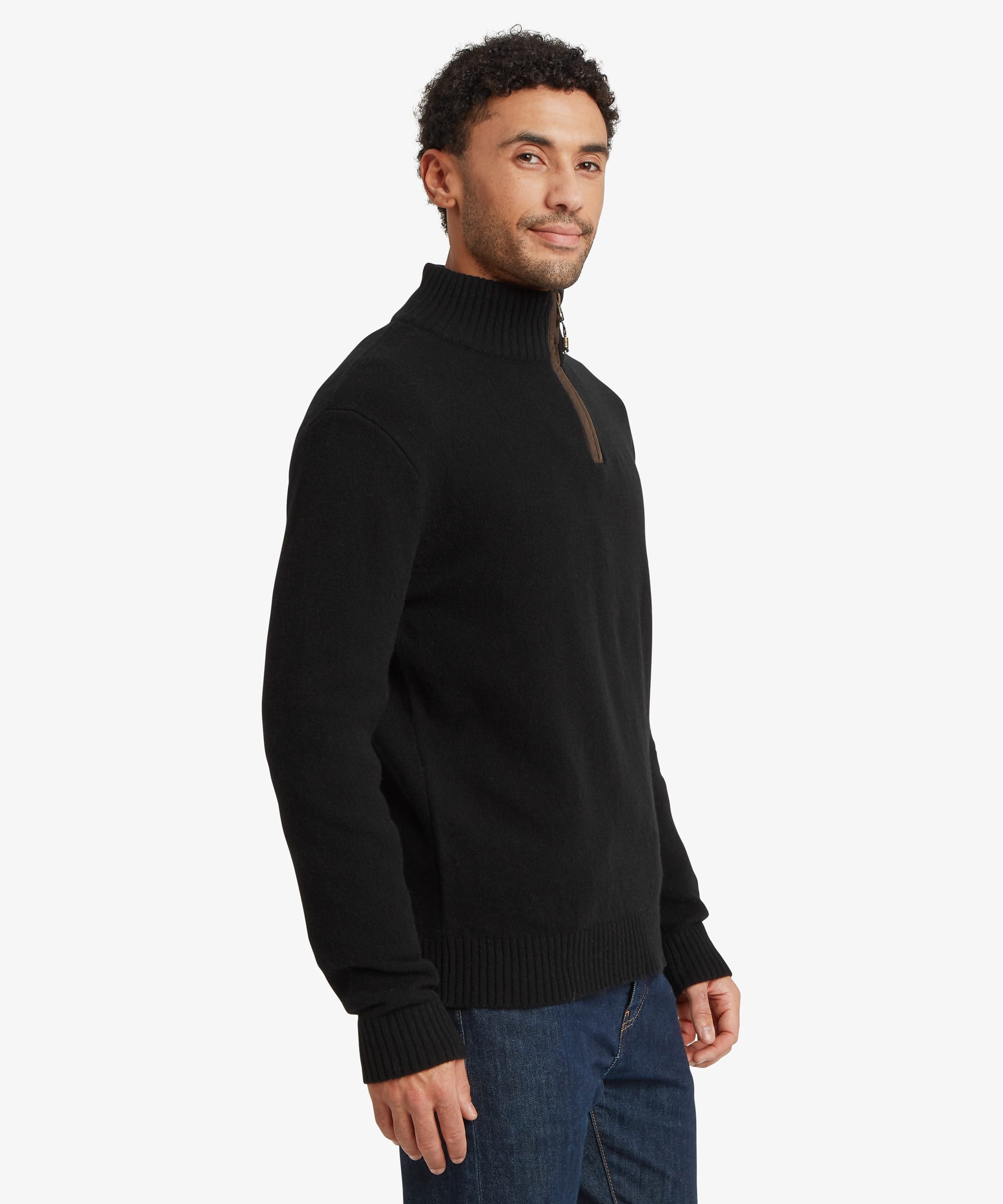 A man wearing a black long-sleeve knit sweater with a mock neck and a small brown leather detail at the collar, paired with blue jeans, photographed against a light gray background.