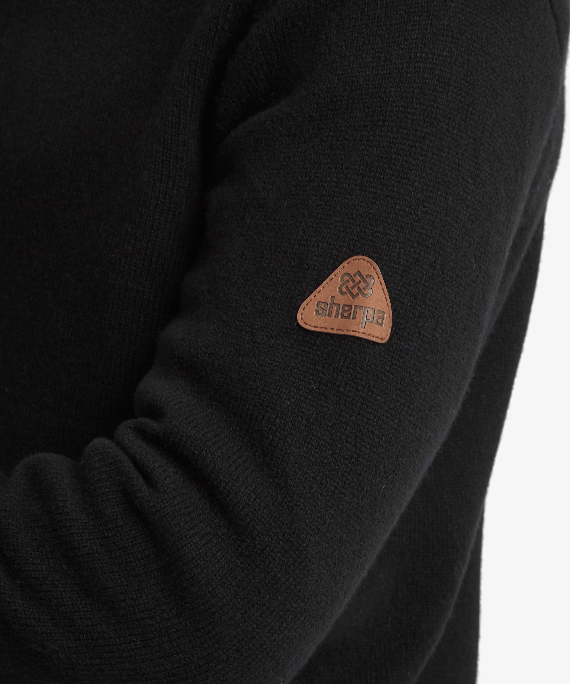 Close-up of a black knit sweater sleeve featuring a small brown triangular patch with the brand name 'sherpa' and logo.