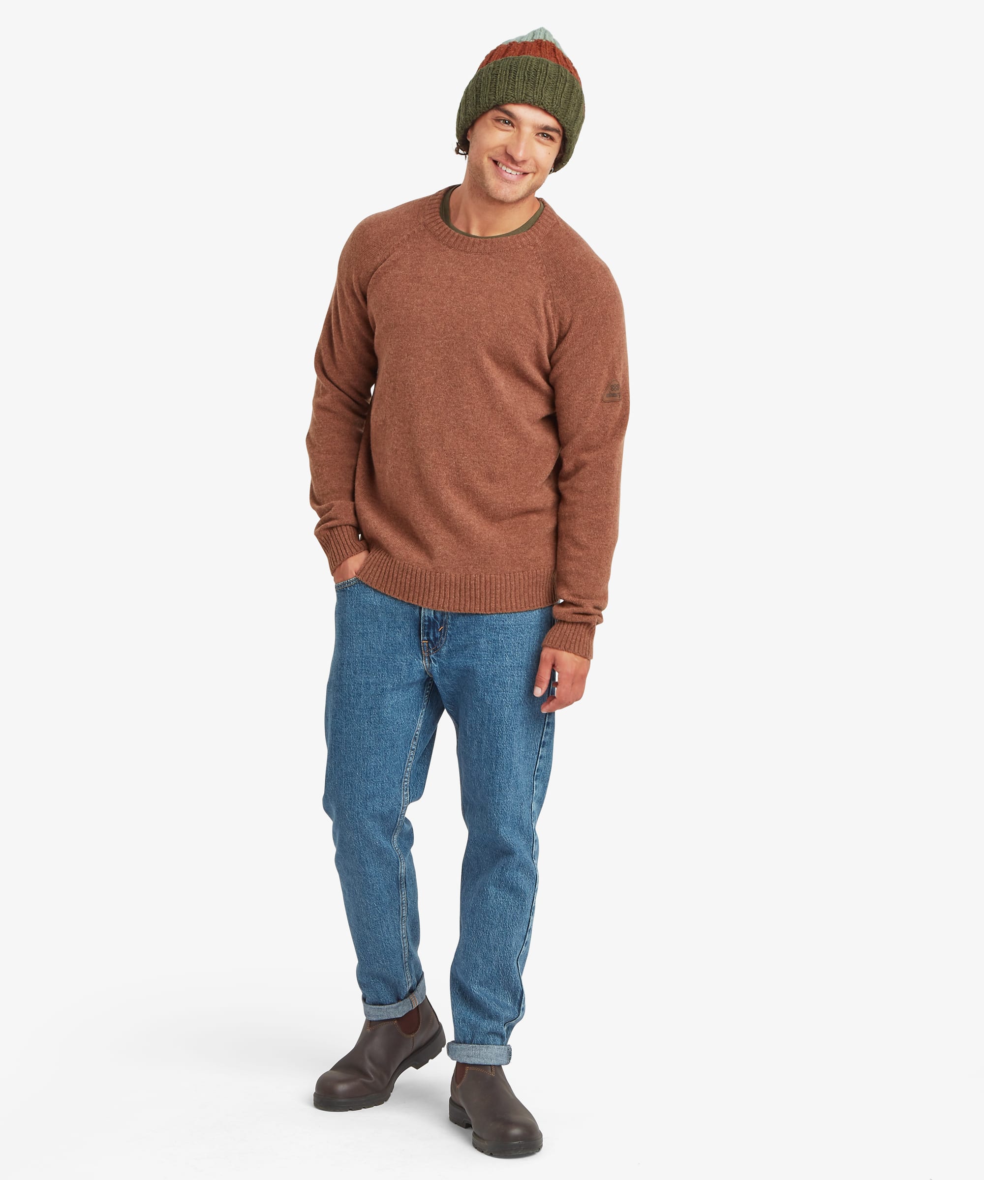 Full-length front view of a male model wearing the Sherpa Kangtega Crew Sweater in brown with blue jeans and a green striped knit beanie. The regular fit, crew neck and ribbed hem and cuffs are clearly visible.