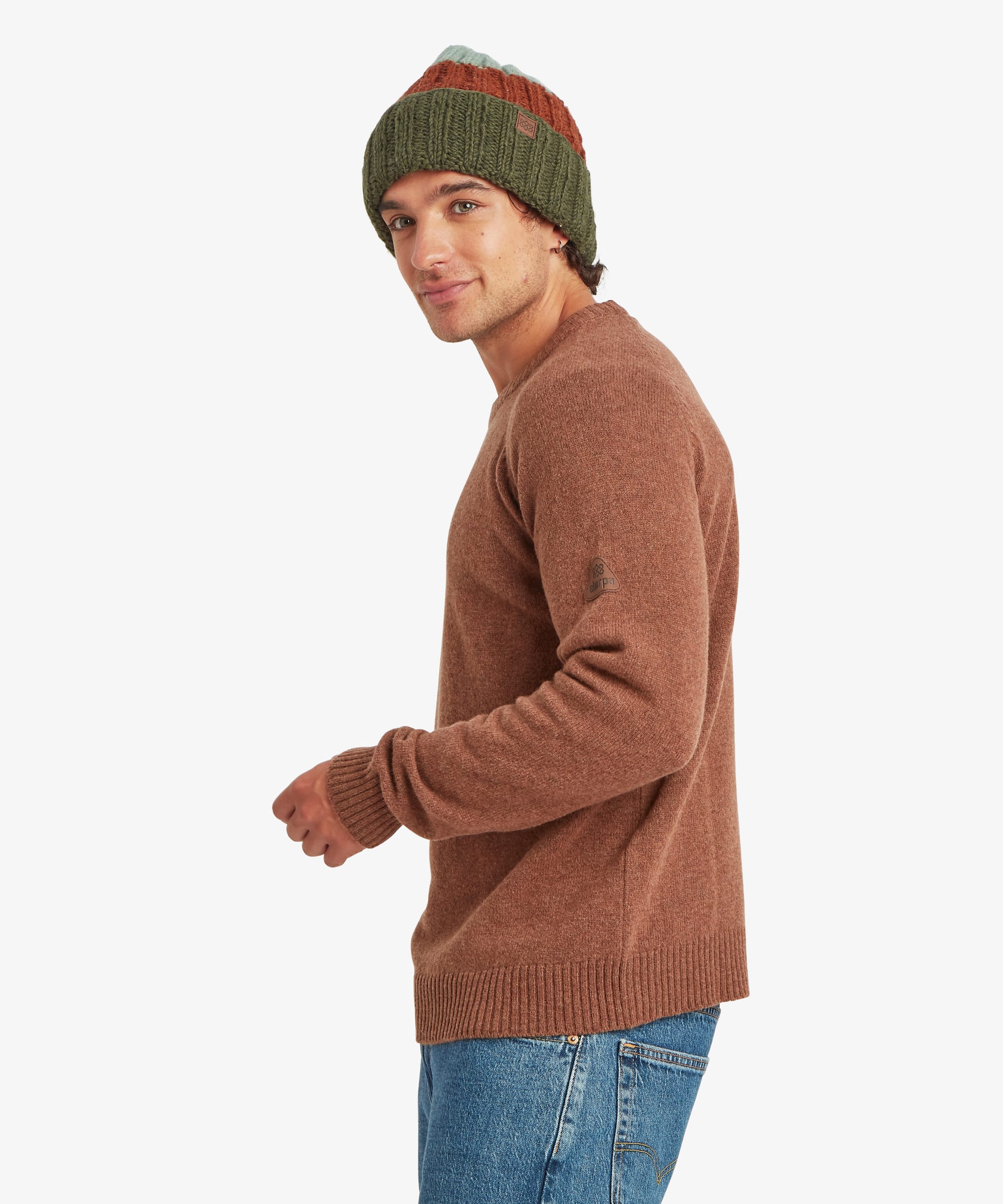 Side profile of a male model wearing the Sherpa Kangtega Crew Sweater in brown with blue jeans and a green striped knit beanie, highlighting the regular fit, crew neckline and straight hem.