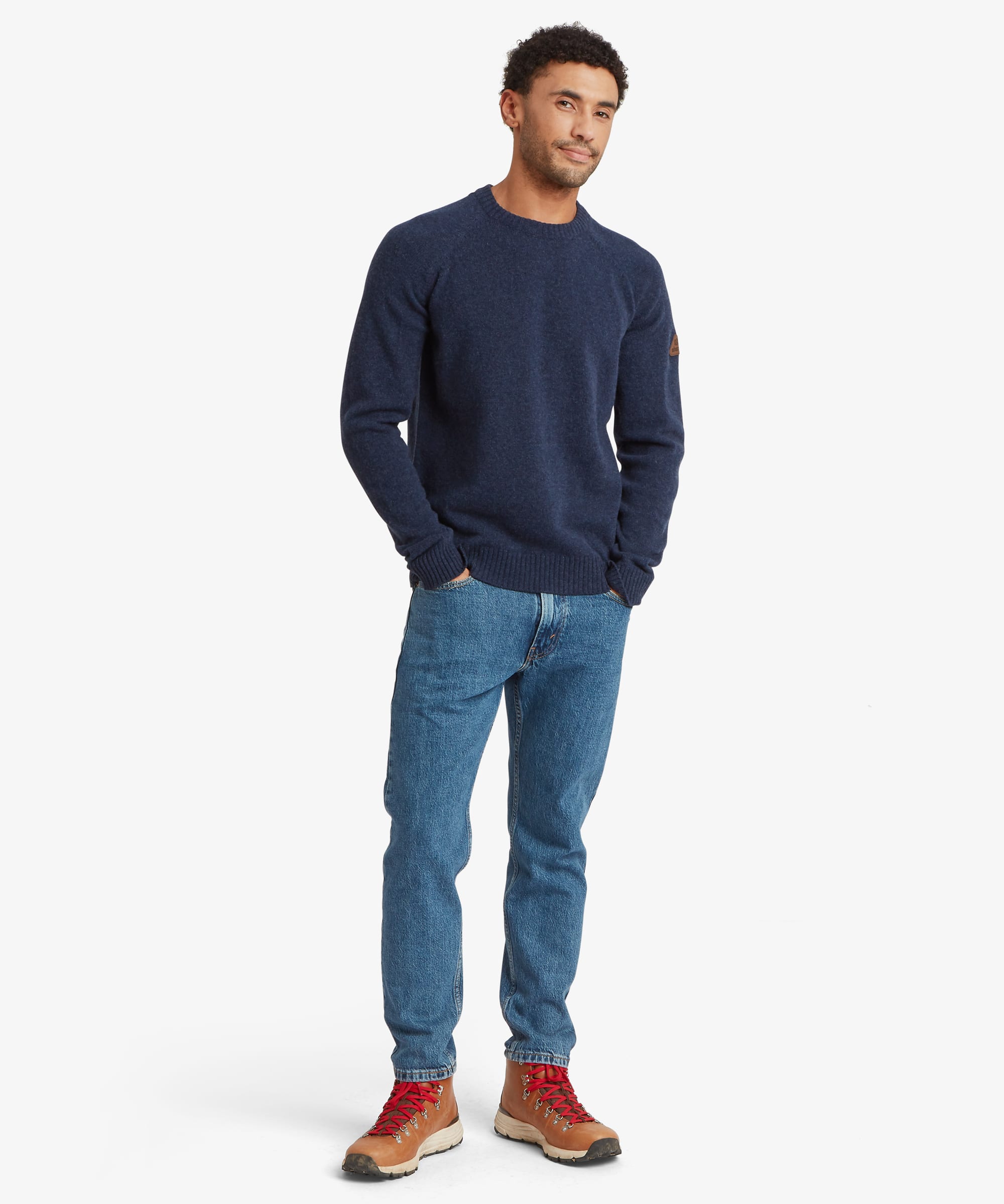 A man wearing a navy blue crewneck sweater, blue jeans, and brown hiking boots with red laces, standing against a light gray studio background.
