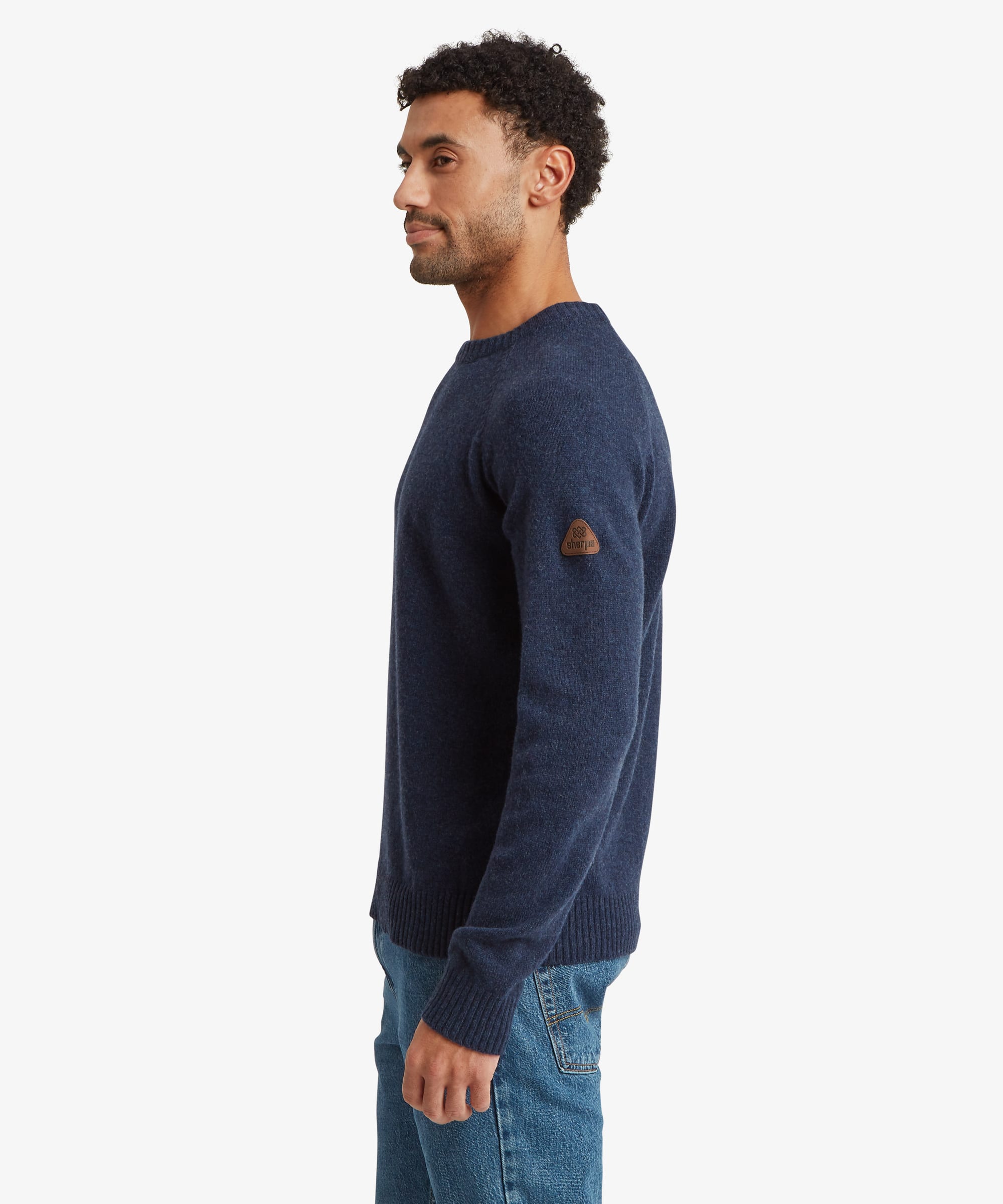 A man shown in profile wearing a navy blue knit sweater with a small brown patch on the upper sleeve, paired with blue jeans, against a plain light gray background.