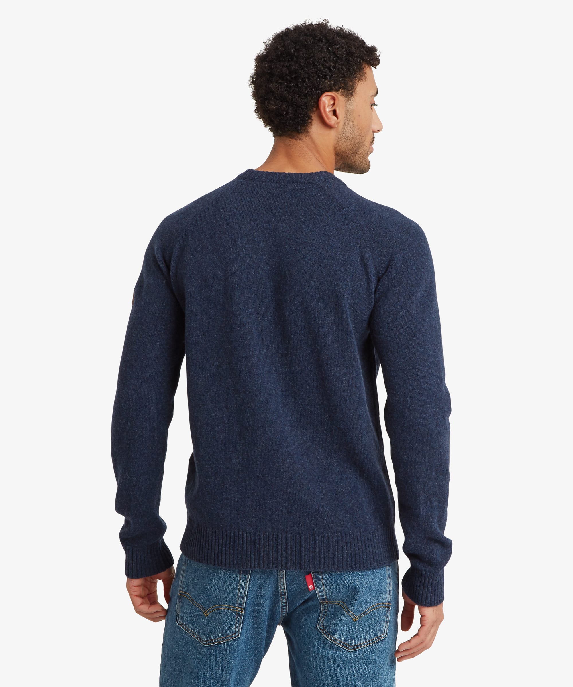 A man viewed from the back wearing a navy blue knit sweater and blue jeans against a light gray studio background. The sweater features ribbing at the cuff, collar and hem.