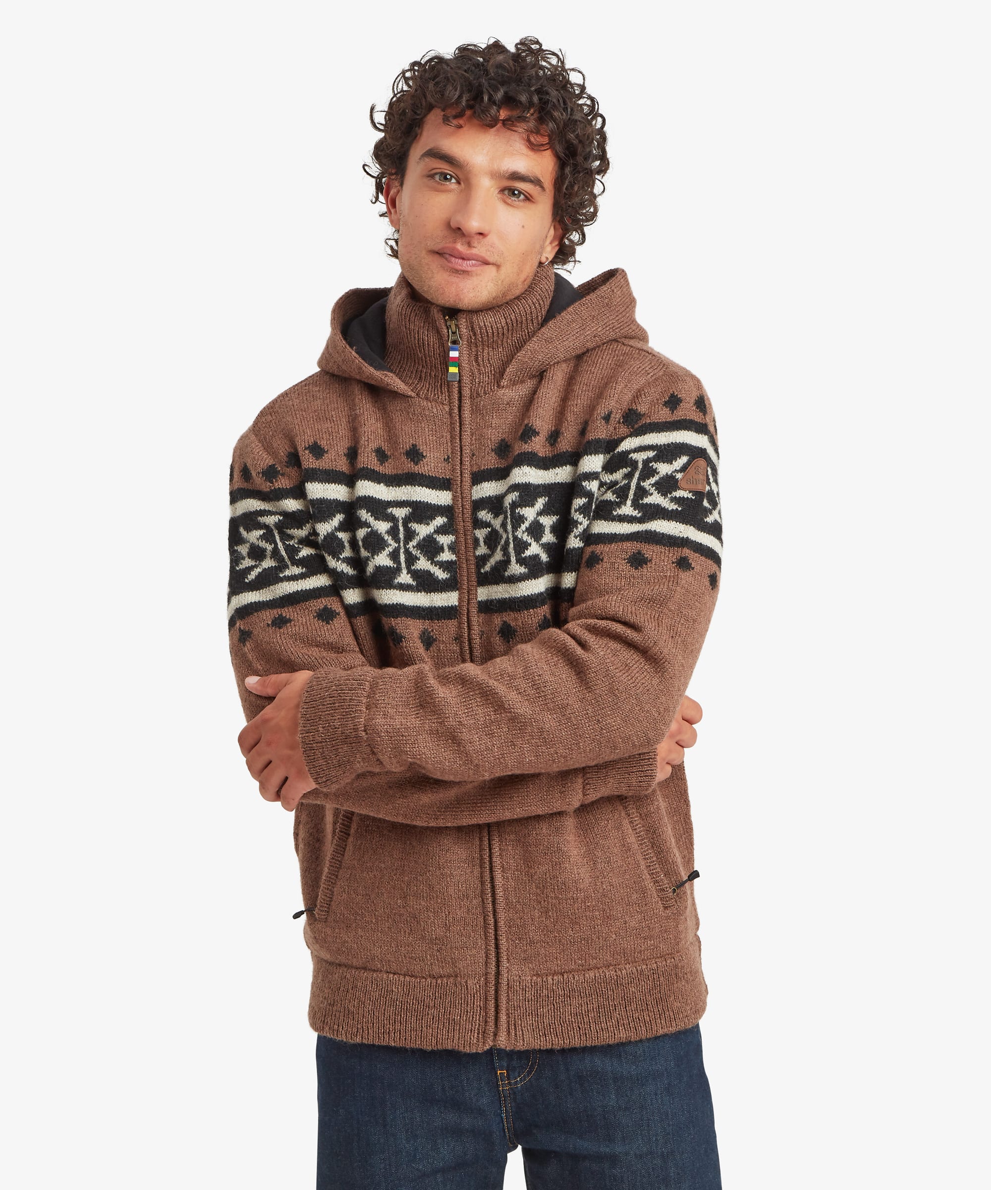 A man wearing a brown zip-up hoodie with a black and white geometric pattern across the chest and shoulders, paired with dark blue jeans, against a light gray background. The sweater features a full zip with a prayer flag zip puller a thick hood.