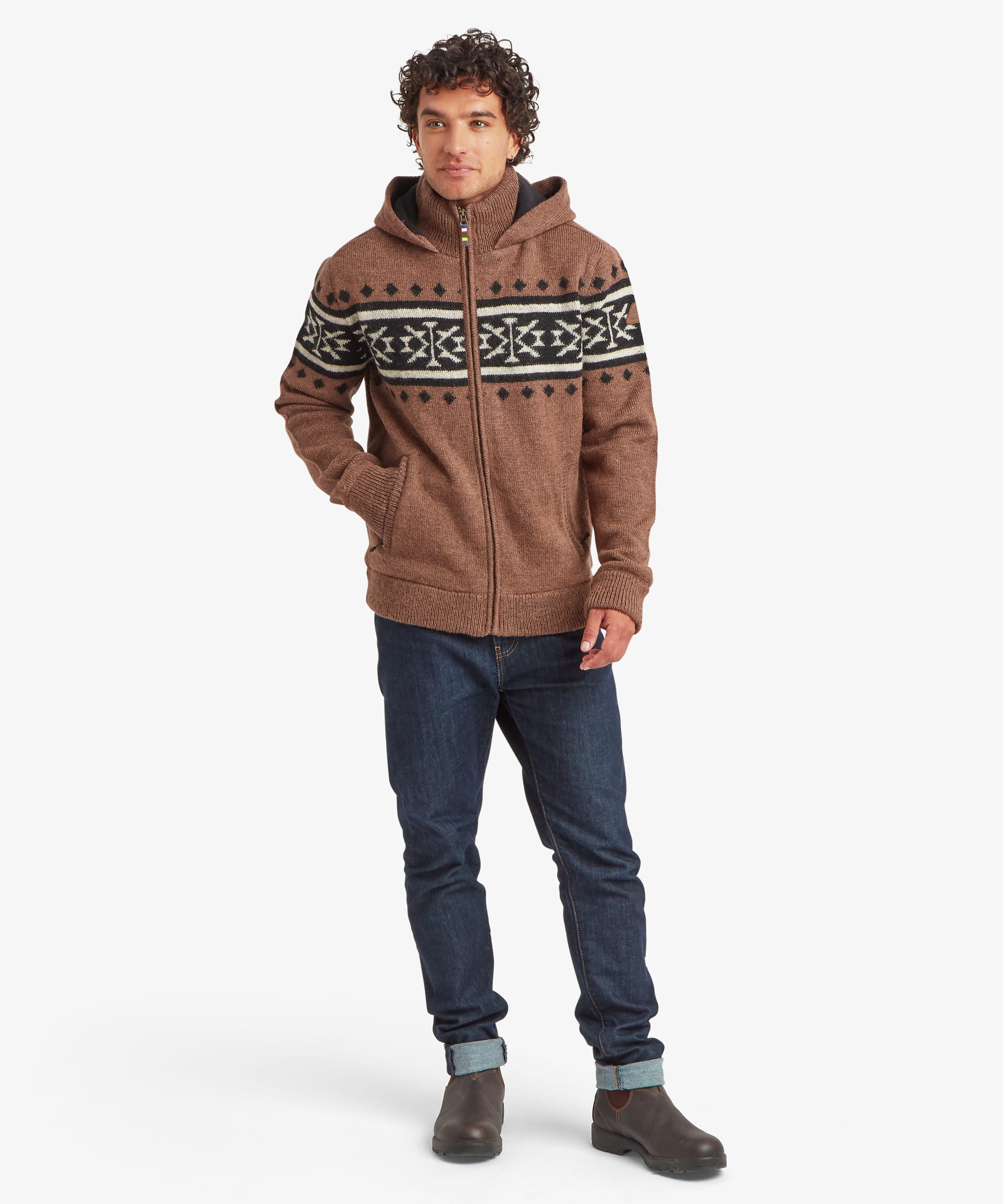 A man wearing a brown knitted zip-up hoodie with a black and white Fair Isle pattern across the chest and shoulders, paired with dark blue jeans and brown boots, standing against a light gray background.