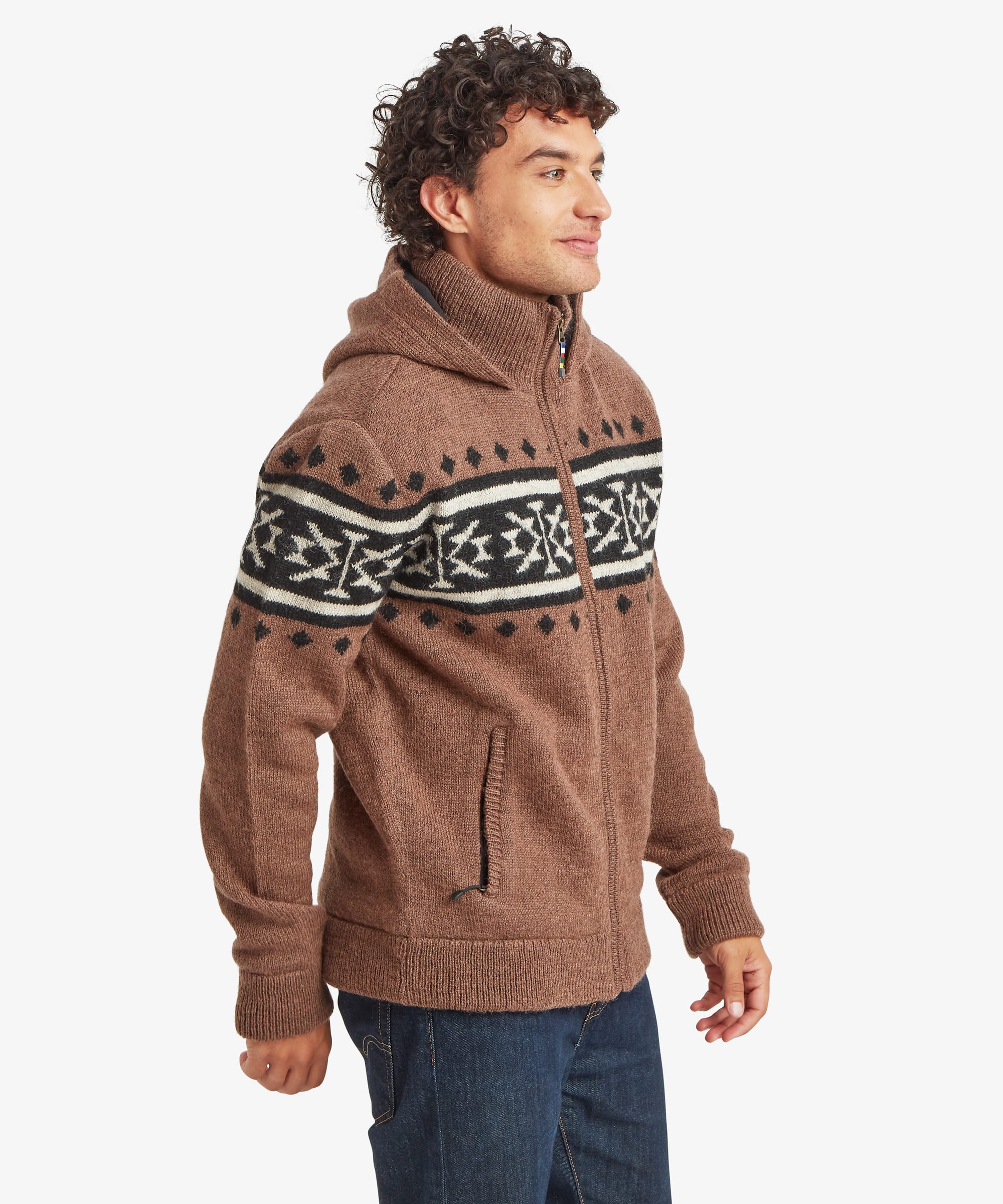A man wearing a brown knit zip-up hoodie with a black and white Nepal inspired pattern across the chest and shoulders, paired with blue jeans, photographed against a light gray background.