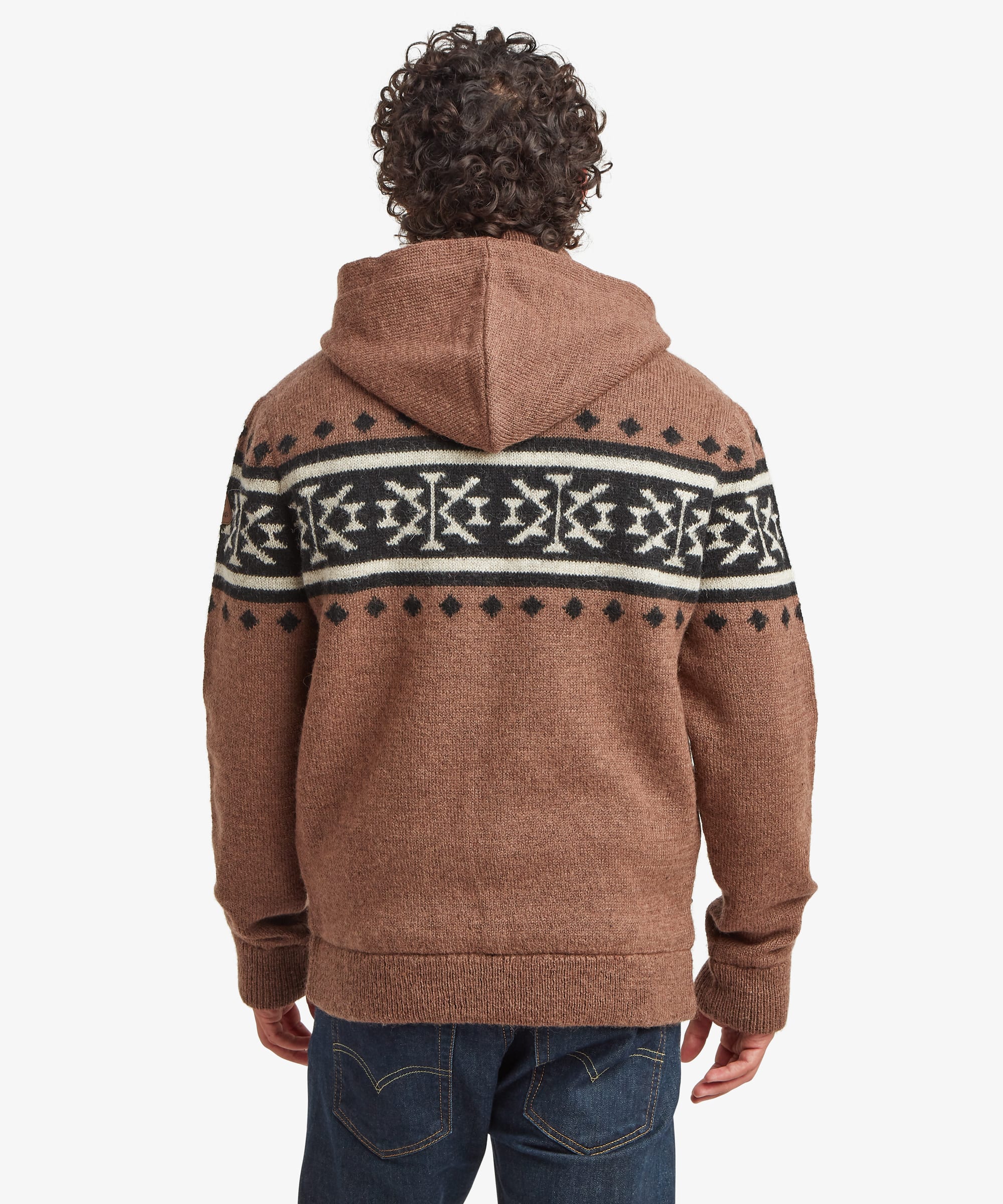 A man viewed from the back wearing a brown hooded knitted sweater with a black and white geometric pattern across the shoulders and upper back, paired with blue jeans, against a light gray studio background.
