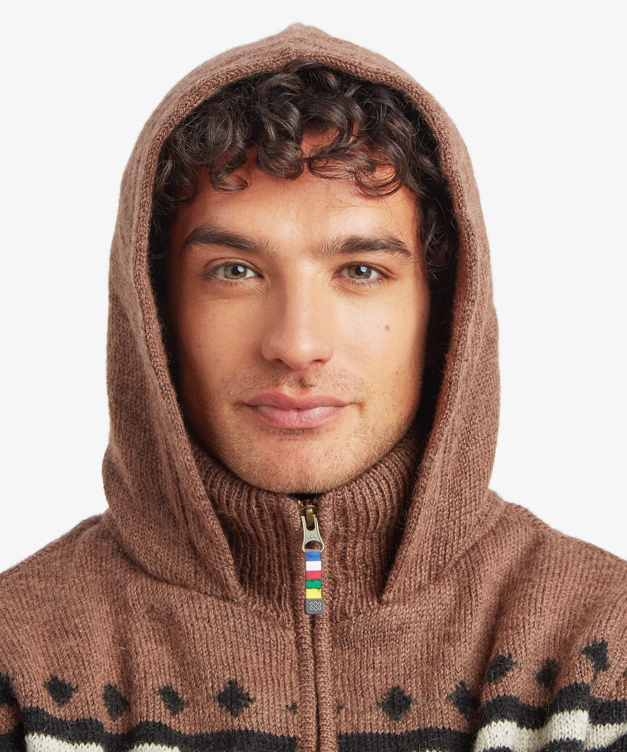 A close-up of a man wearing a brown knit zip-up hoodie with a high ribbed collar and hood, featuring a small multicolor zipper pull and black and white patterned knit details at the chest, against a light gray background.