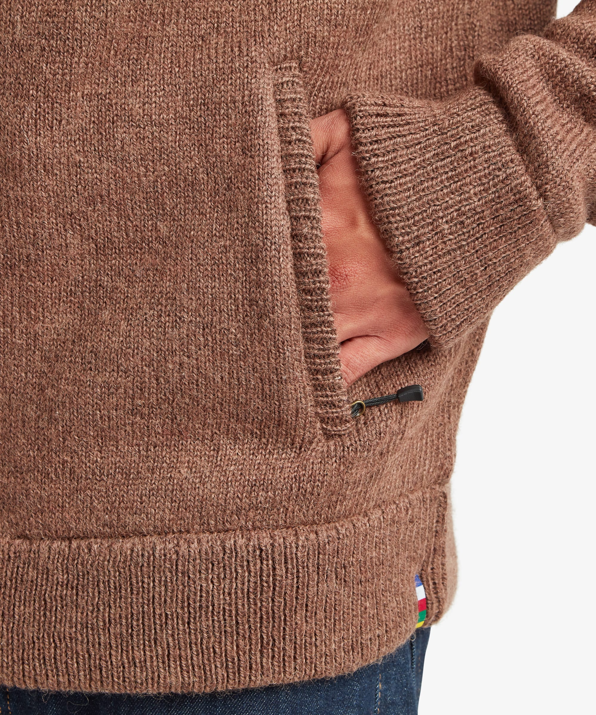Close-up of a brown knit sweater with a side pocket and a hand tucked inside, showing the ribbed cuff and hem against a light gray background.