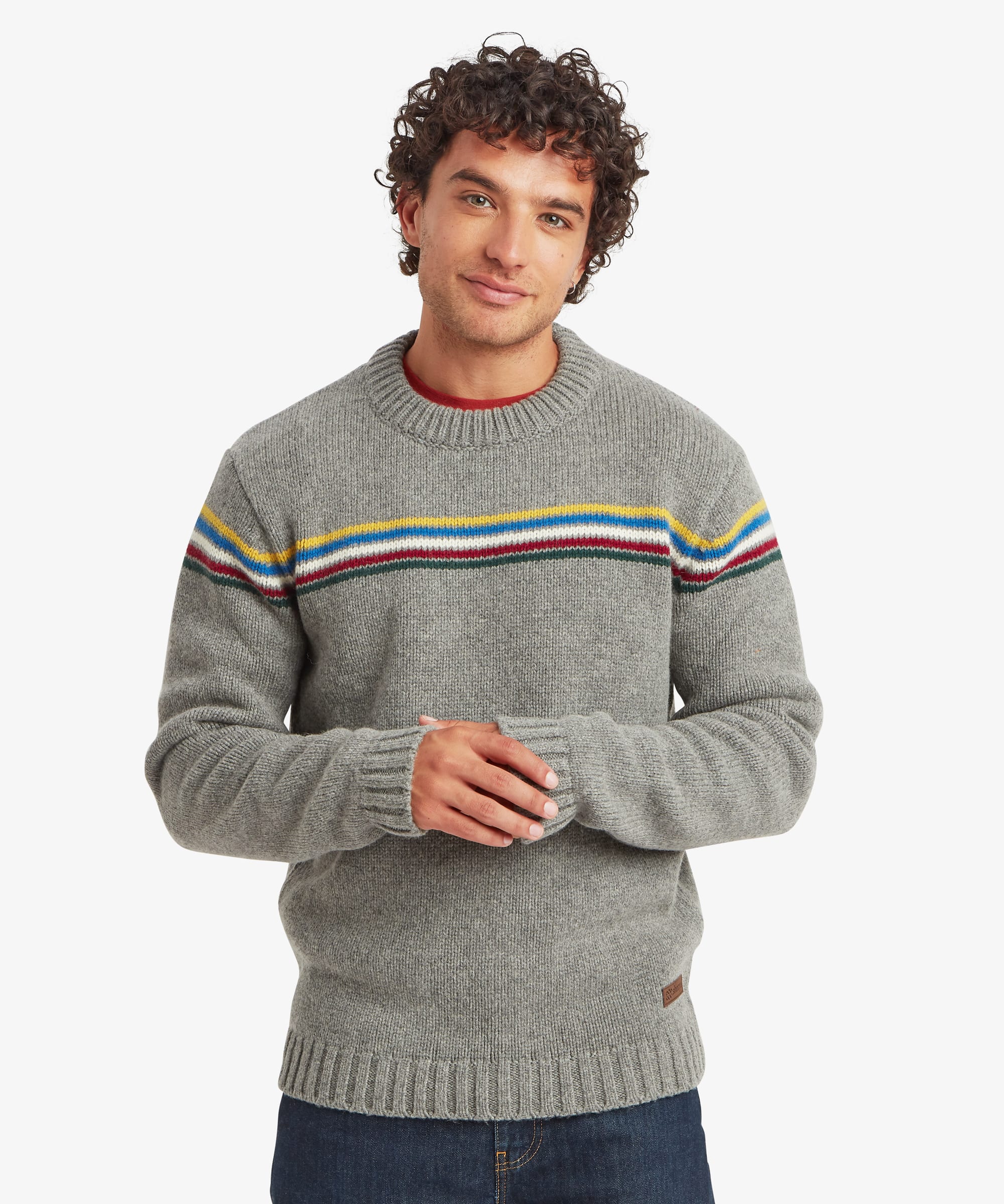 Front view of a male model wearing the Sherpa Mahesh Sweater in grey with dark blue jeans. The knitted crew neck sweater features colourful horizontal chest stripes, long sleeves, ribbed cuffs and hem, and a regular fit.