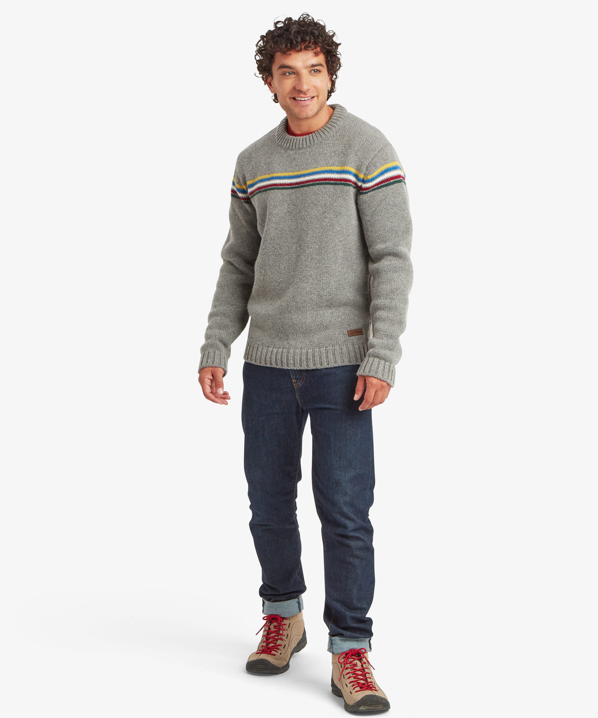 Full-length front view of a male model wearing the Sherpa Mahesh Sweater in grey with dark blue jeans, highlighting the classic crew neckline, colourful striped chest detail and relaxed regular fit.