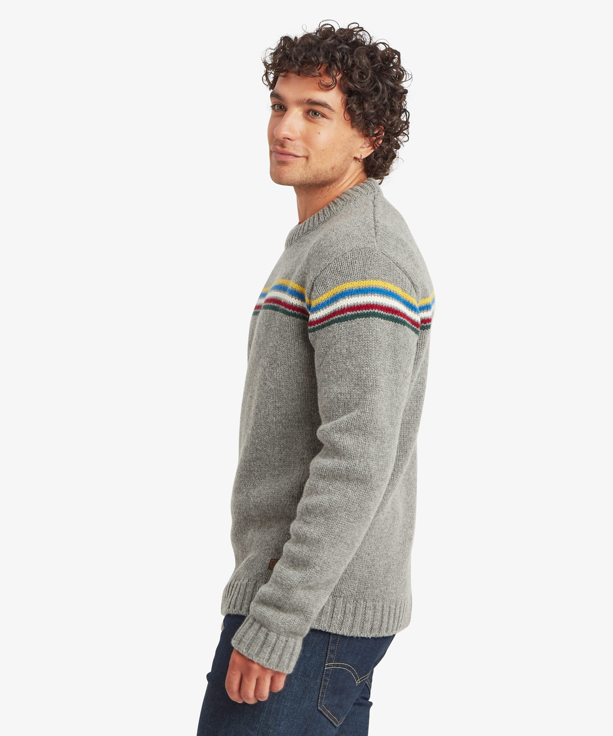 Side profile of a male model wearing the Sherpa Mahesh Sweater in grey with dark blue jeans, showing the regular fit, ribbed cuffs and hem, and colourful chest stripe design.