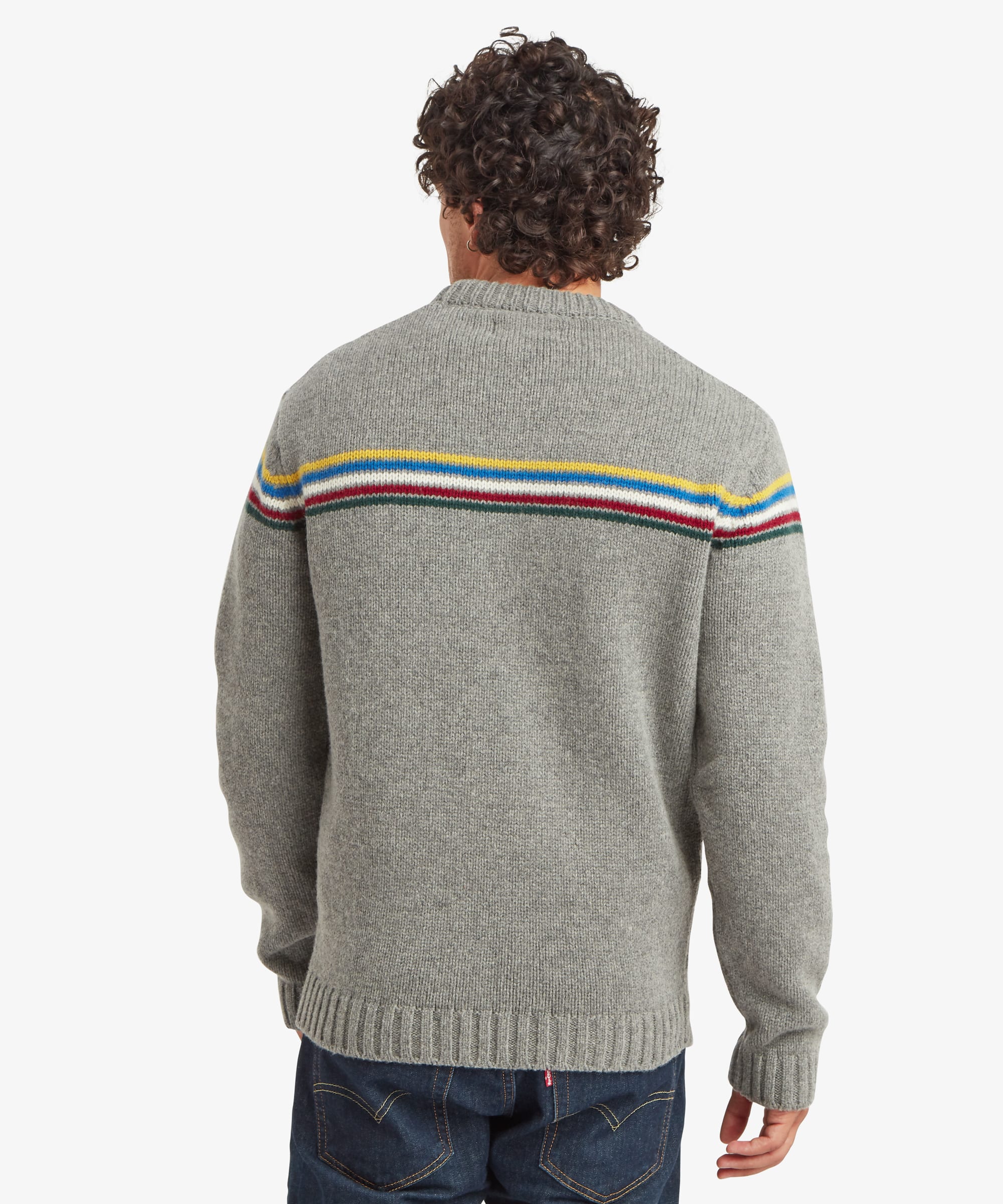 Rear view of a male model wearing the Sherpa Mahesh Sweater in grey with dark blue jeans, displaying the colourful stripe continuing across the upper back, long sleeves and ribbed hem.