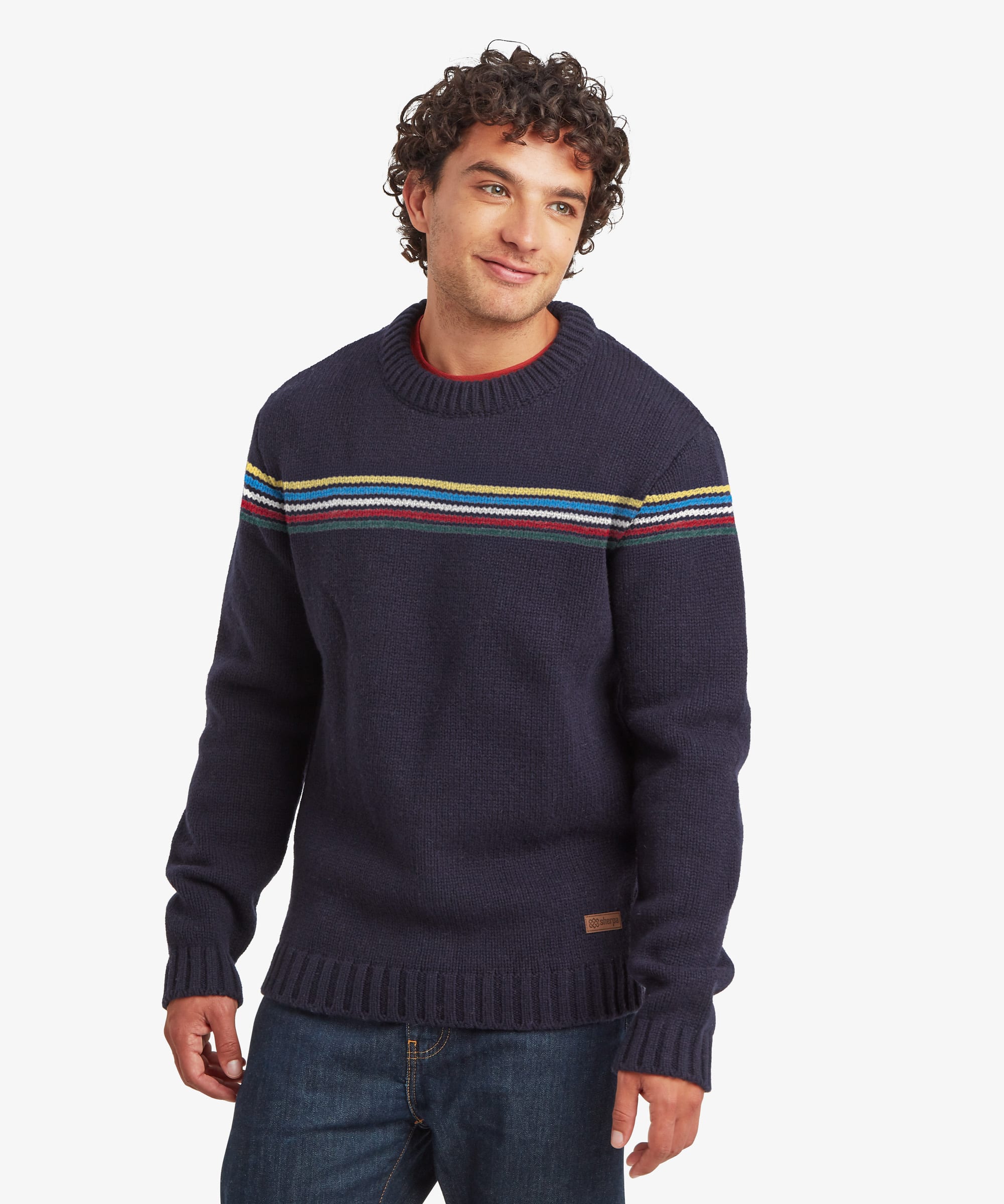 A man wearing a navy blue knit sweater with horizontal stripes in red, yellow, and green across the chest, paired with blue jeans, against a light gray background. The sweater features ribbed details around the hem, collar and cuffs.