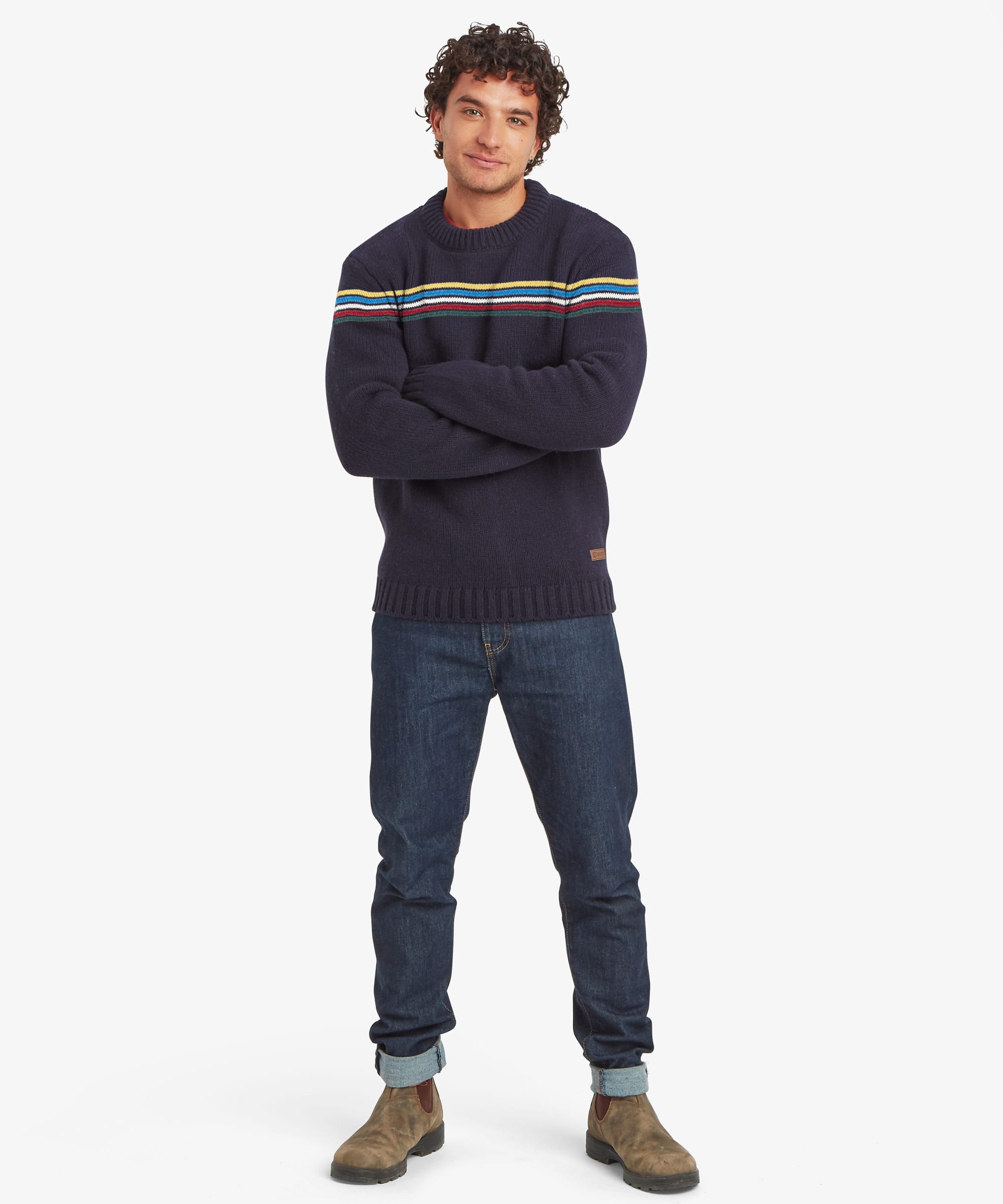 Full-length front view of a male model wearing the Sherpa Mahesh Sweater in blue with dark blue jeans. The knitted crew neck sweater features horizontal blue and white chest stripes, long sleeves, ribbed cuffs and hem, and a comfortable regular fit.