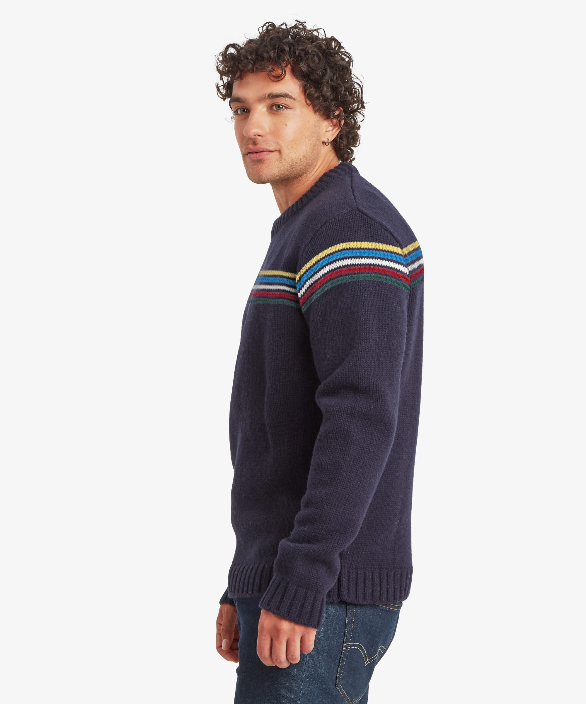 Side profile of a male model wearing the Sherpa Mahesh Sweater in blue with dark blue jeans, highlighting the regular fit, crew neckline, ribbed trims and contrasting striped chest design.