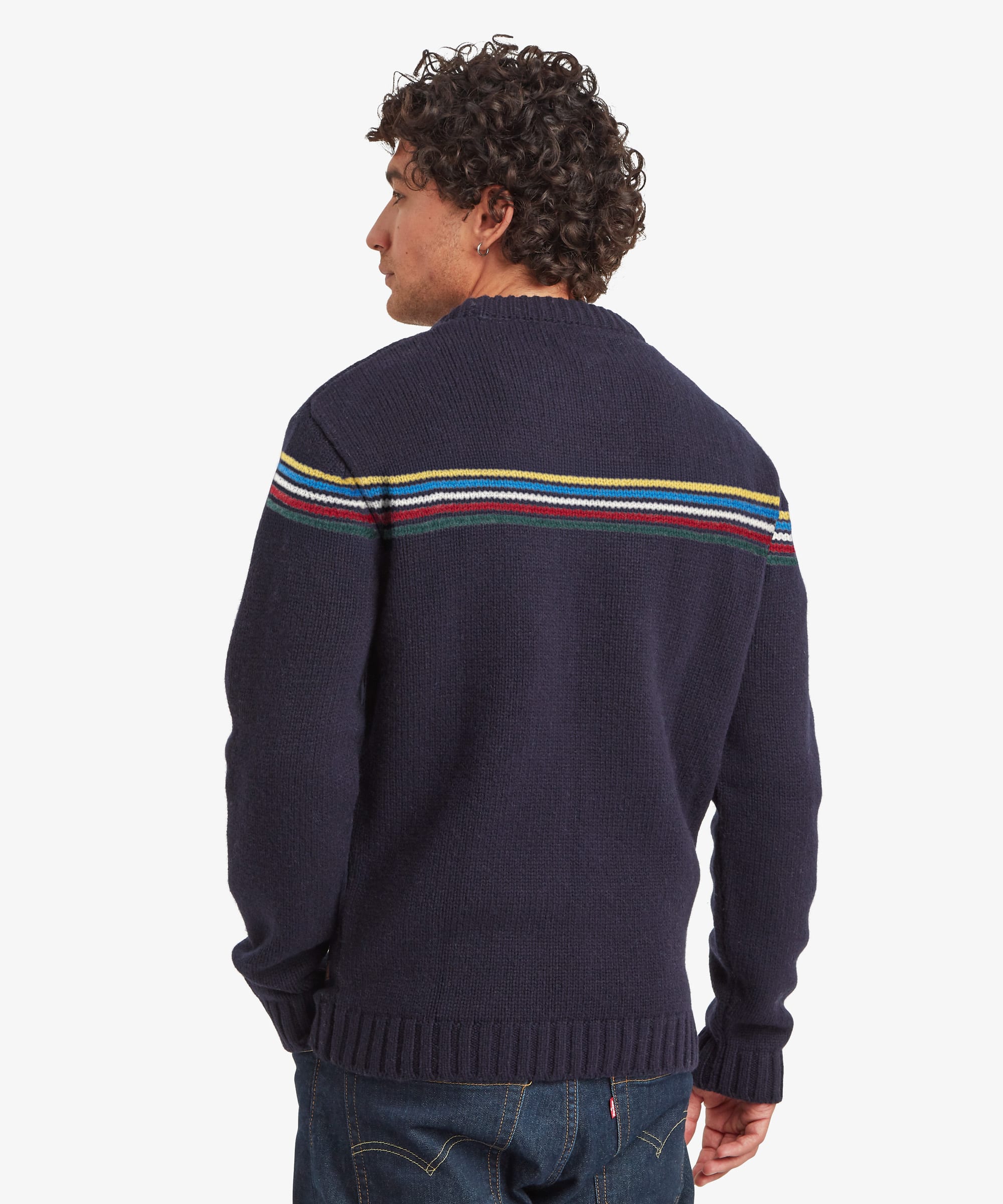 Rear view of a male model wearing the Sherpa Mahesh Sweater in blue with dark blue jeans, showing the striped detail wrapping across the back shoulders, long sleeves and ribbed hem.