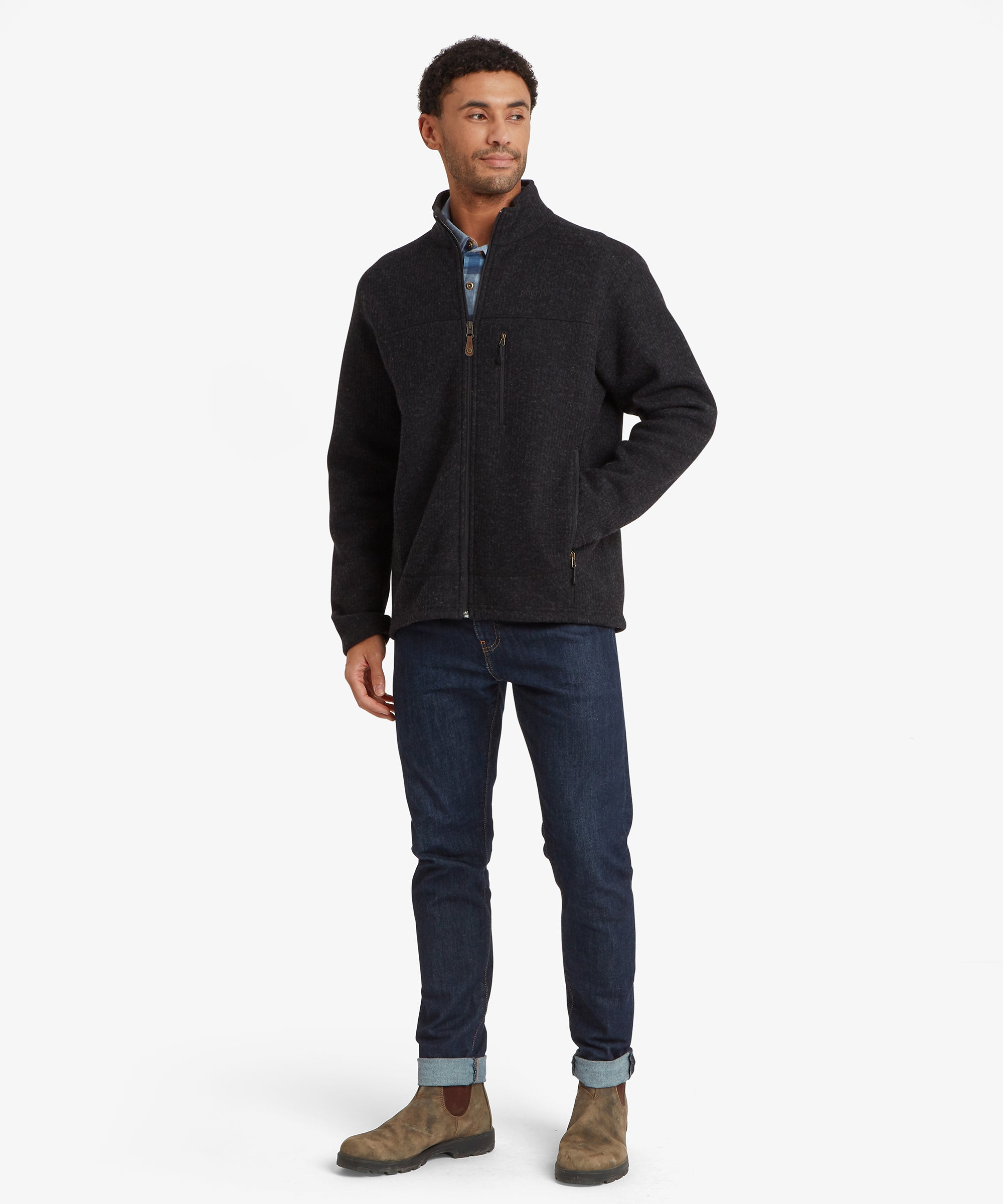 A full length portrait of a man wearing a grey / black woollen fleece jacket and jeans paired with brown boots against a white background. The jacket features multiple pockets including a zipped chest pocket and two and pockets. The model is standing with his left hand in the jacket pocket.