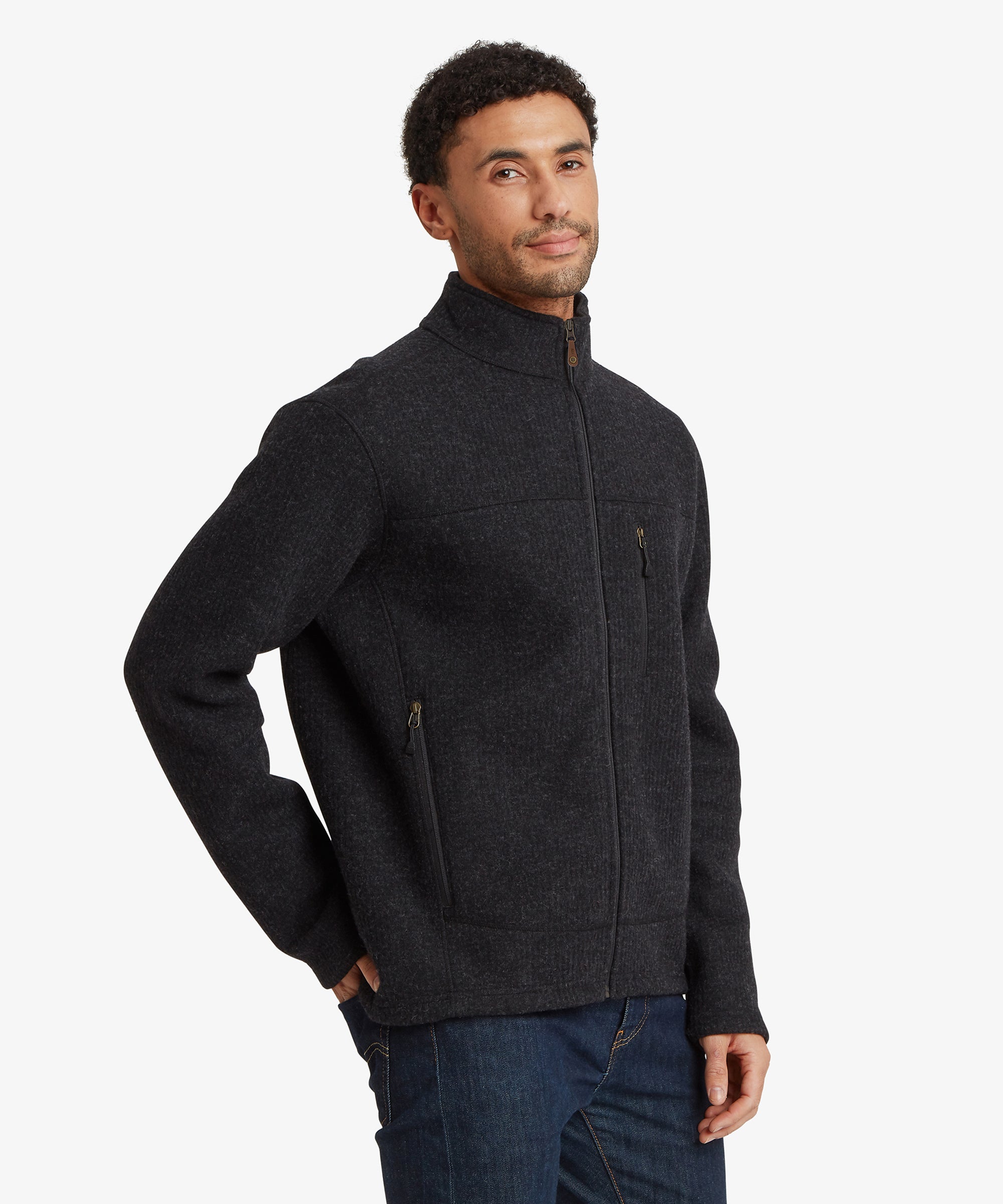 Man wearing a dark gray / black jacket on a white background. The model is looking ahead with his right hand in his back trouser pocket. The jacket is fully zipped to the collar. The jacket features multiple pockets including two hand pockets and a chest pocket.