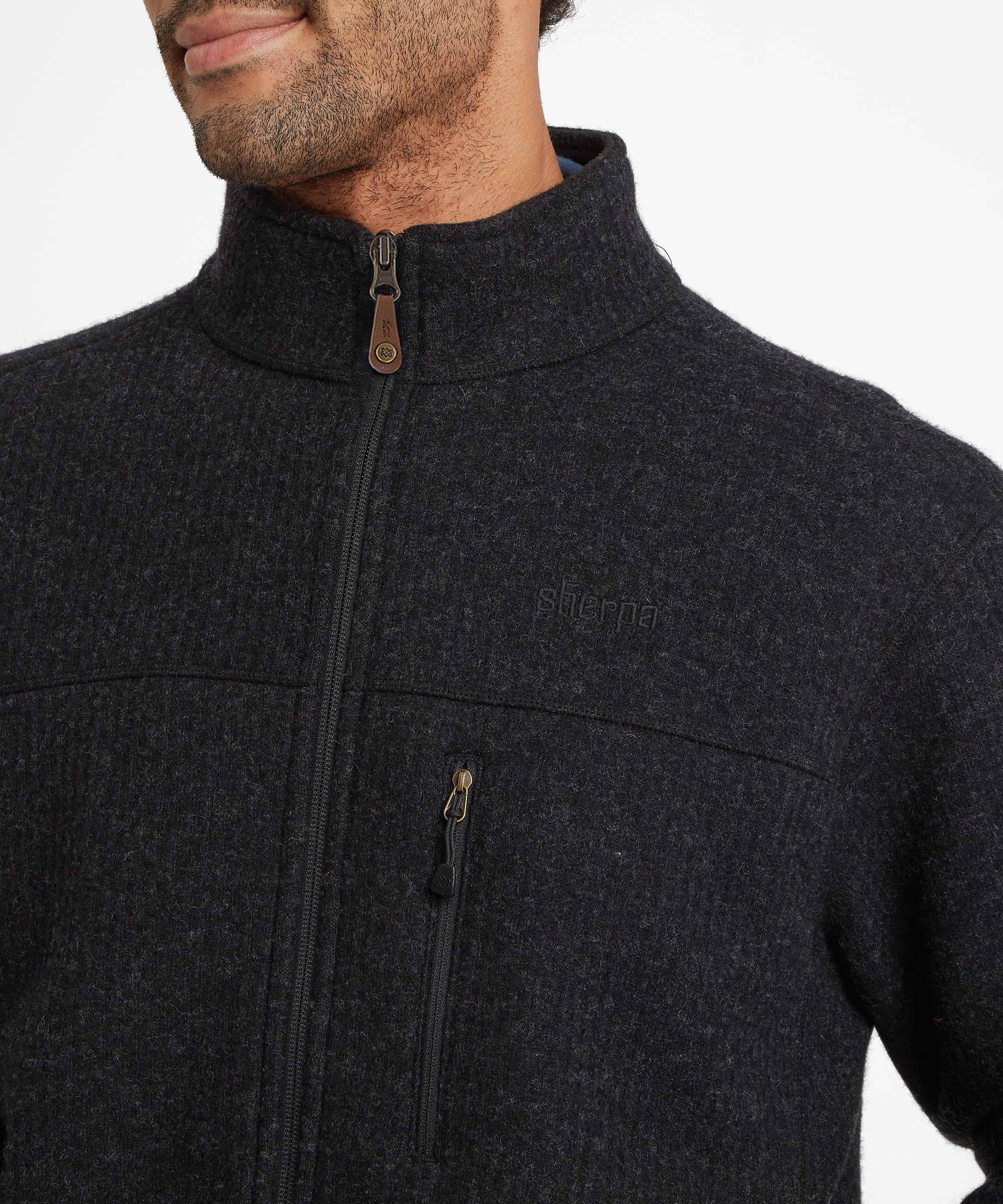 A close up of a man modeling a dark gray fleece jacket with the Sherpa Adventure Gear brand logo embroidered on the chest standing against a white background. The fleece jacket features a full zip, with a faux leather zip puller with an endless knot stud, and a zipped chest pocket with a string puller.