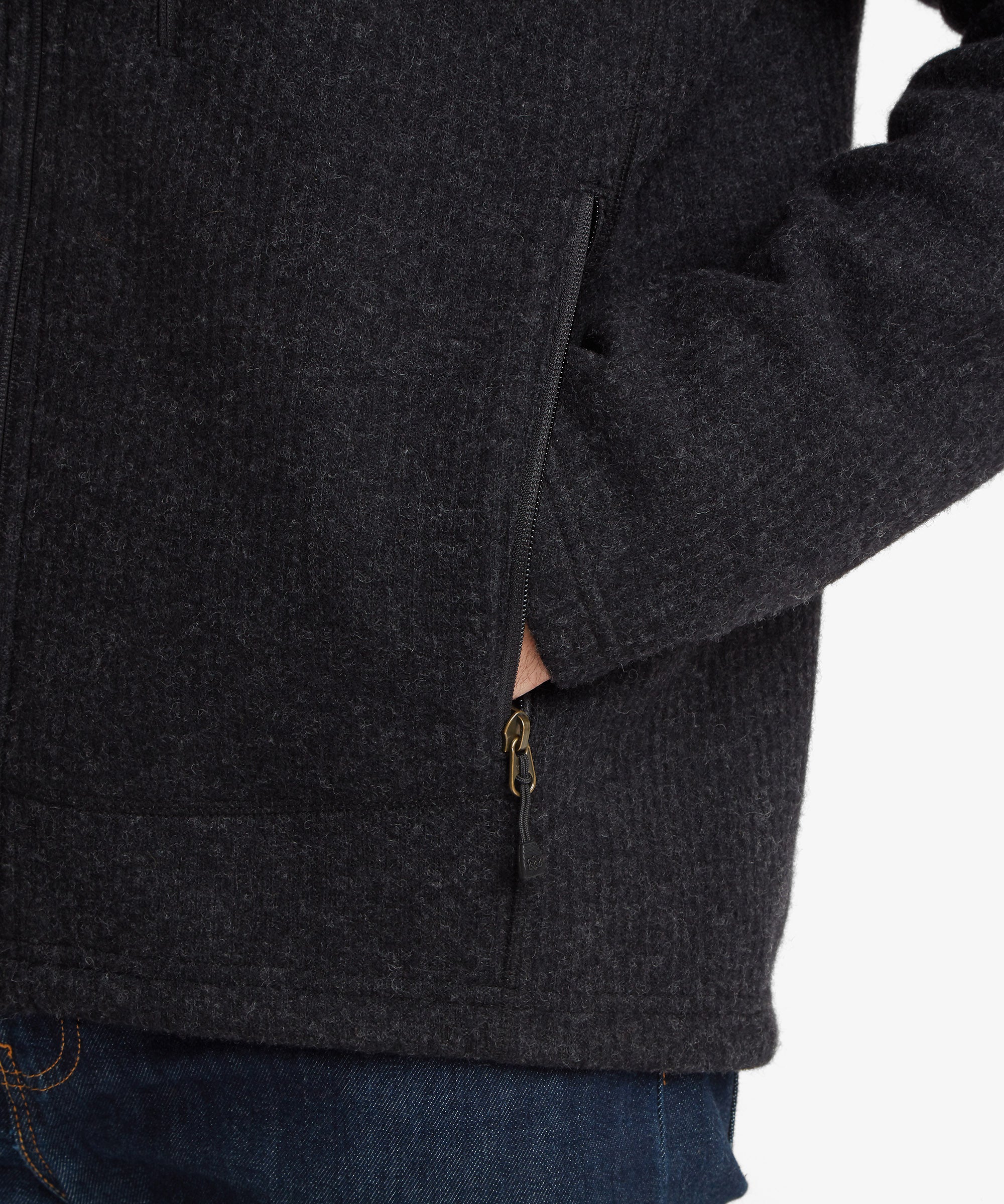 Close-up of a dark charcoal gray zippered woollen, fleece jacket worn by a model, showing the side pocket with a hand inside and a brass zipper pull, paired with blue jeans against a white background.